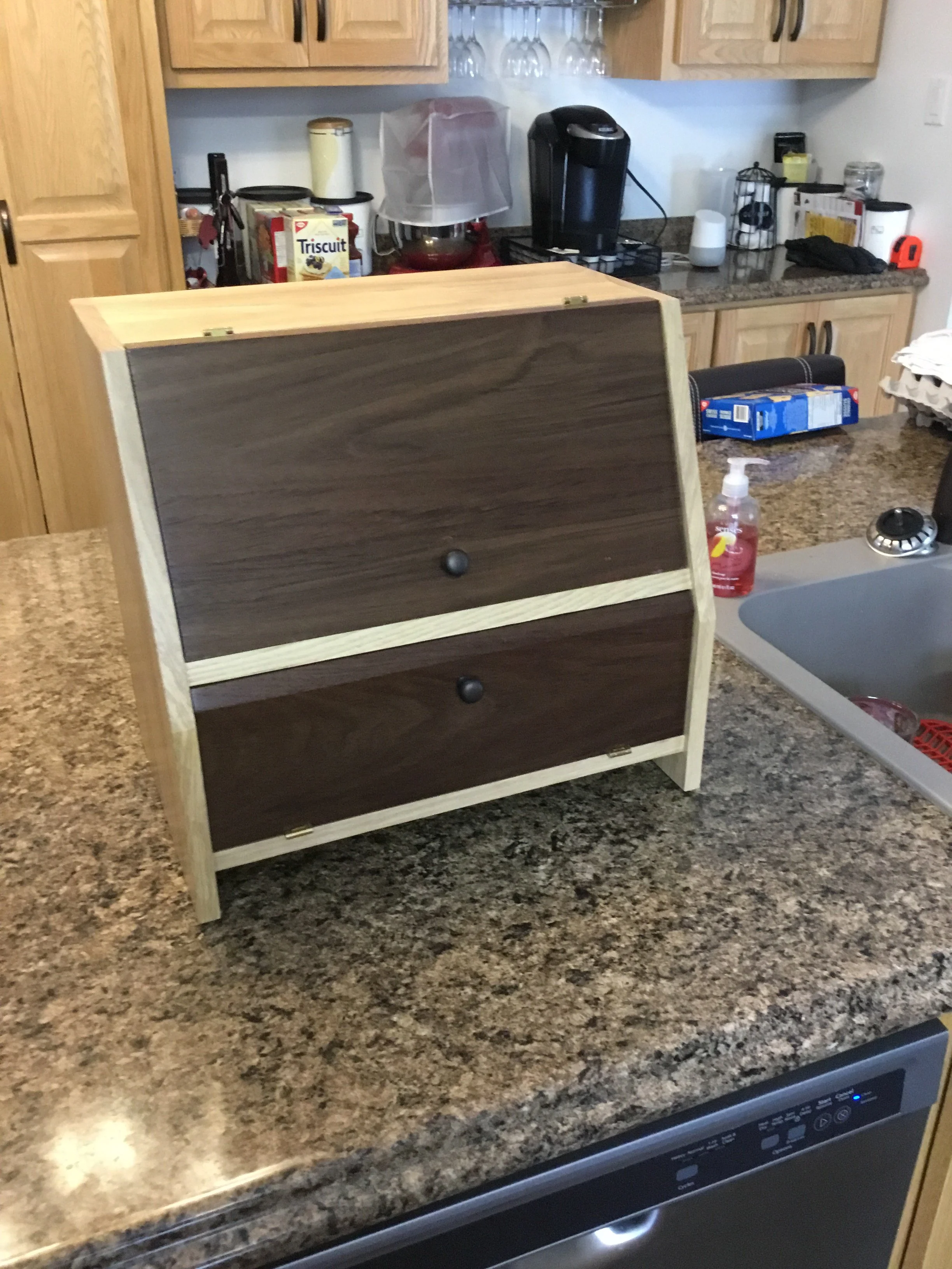 A kitchen countertop with a partially assembled wooden drawer or cabinet piece, various kitchen appliances, and items, including a box of Triscuit crackers and a bottle of hand sanitizer.