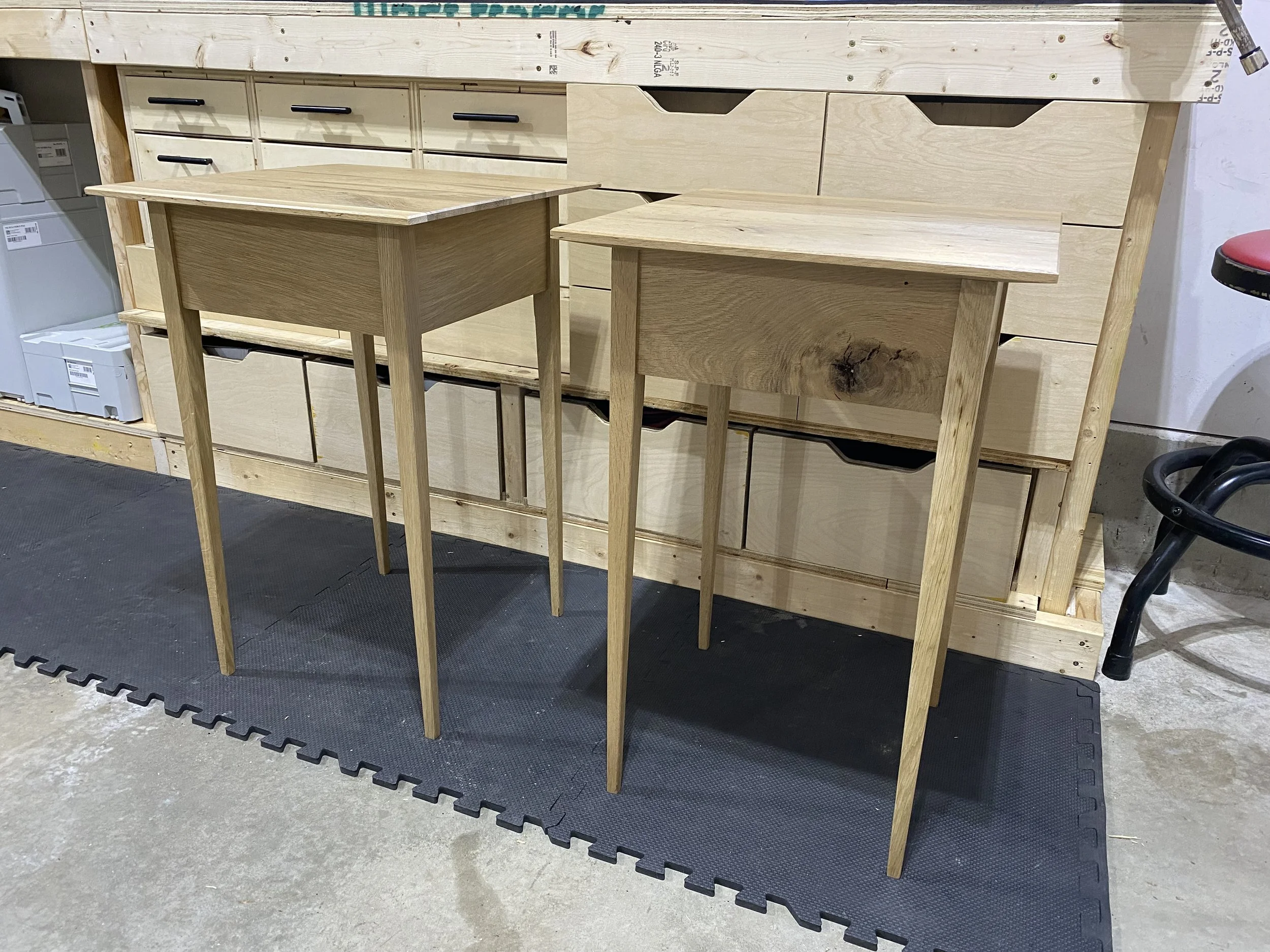Two unfinished wooden tables in front of a large wood piece with drawers, in a workshop setting.