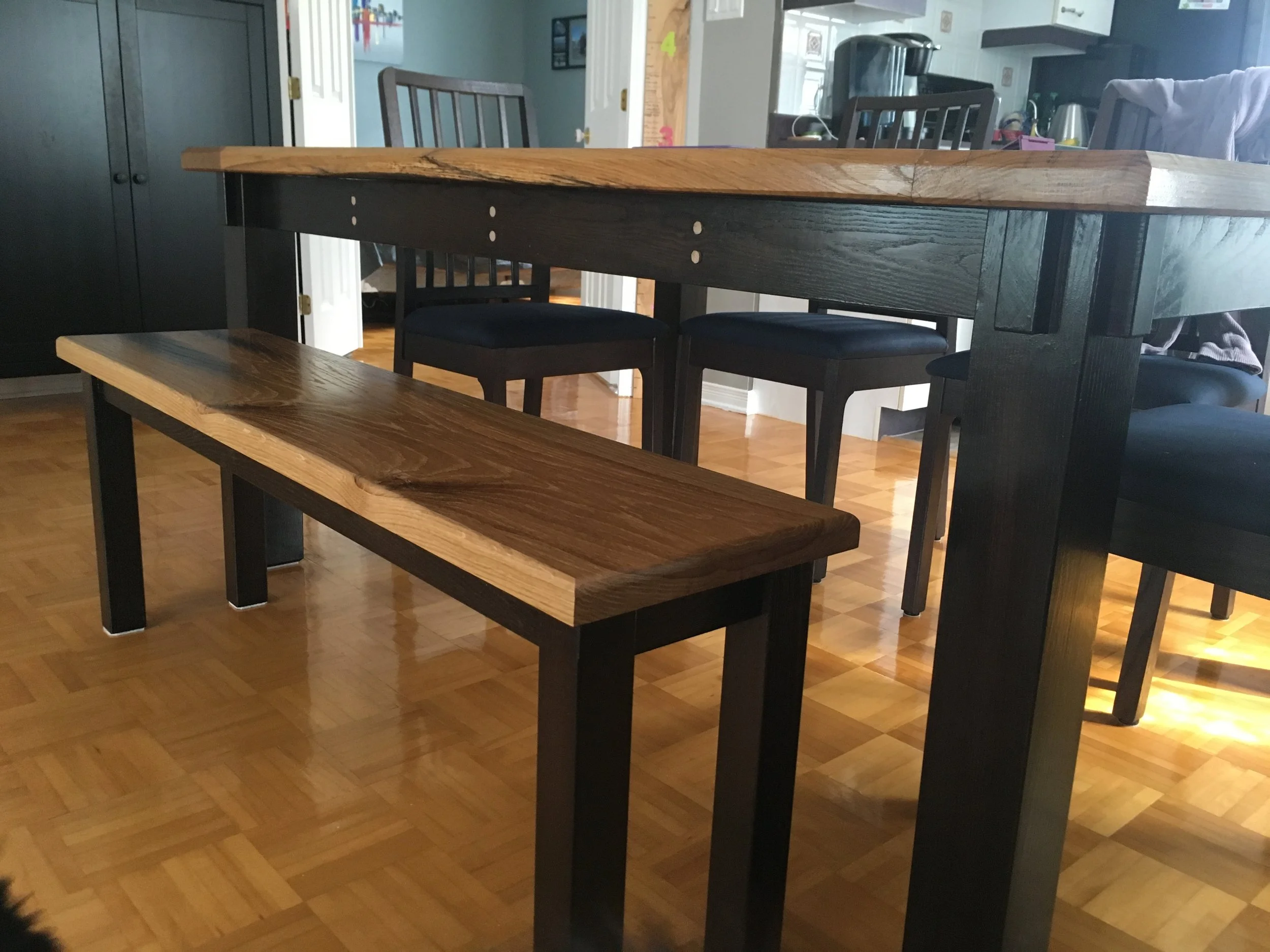 A dining area with a wooden table and black chairs in a home kitchen. The background shows a kitchen counter with appliances and a living room with a couch and various household items.