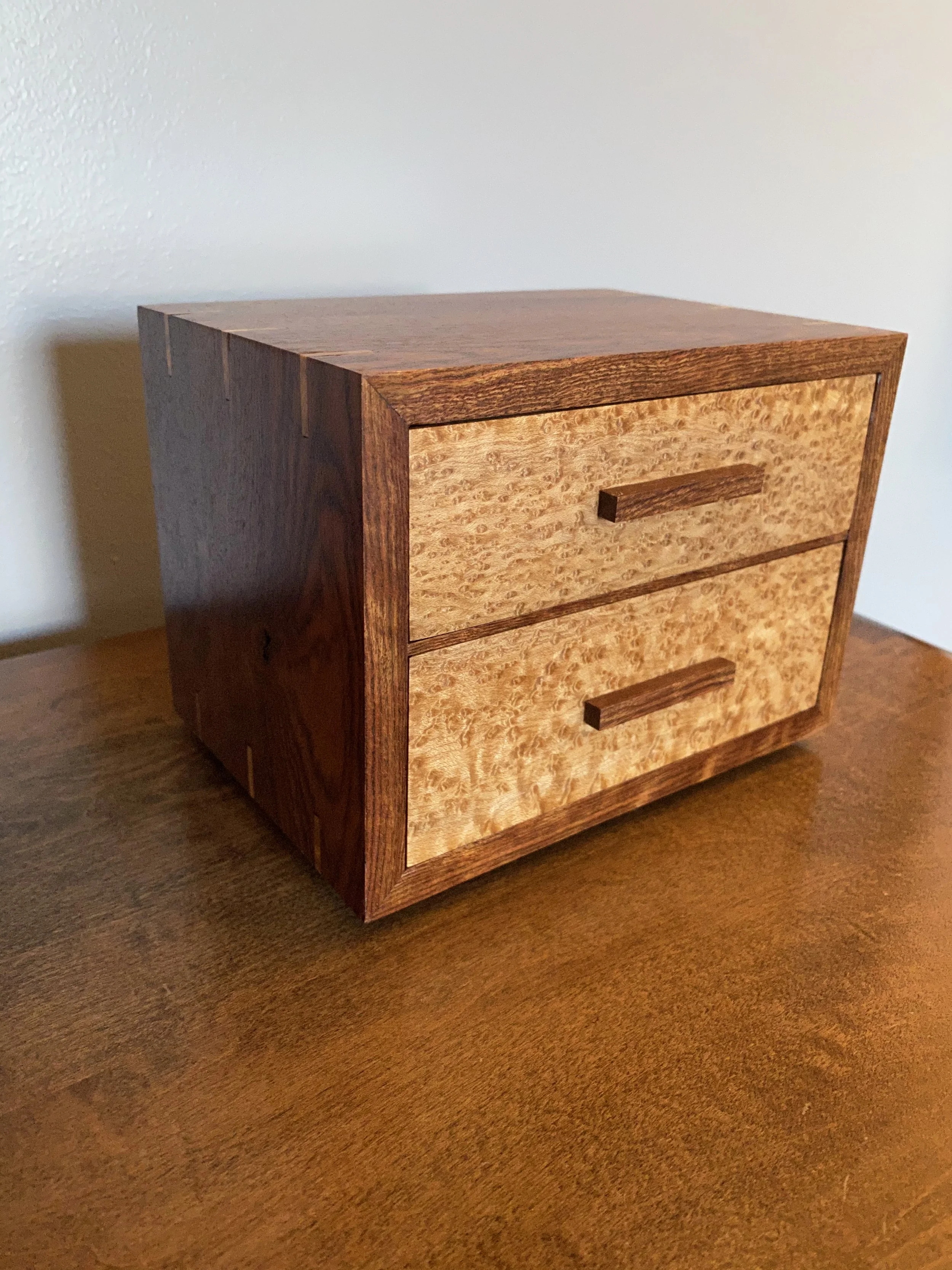 A wooden nightstand with three drawers and horizontal handles, placed on a wooden floor against a plain white wall.