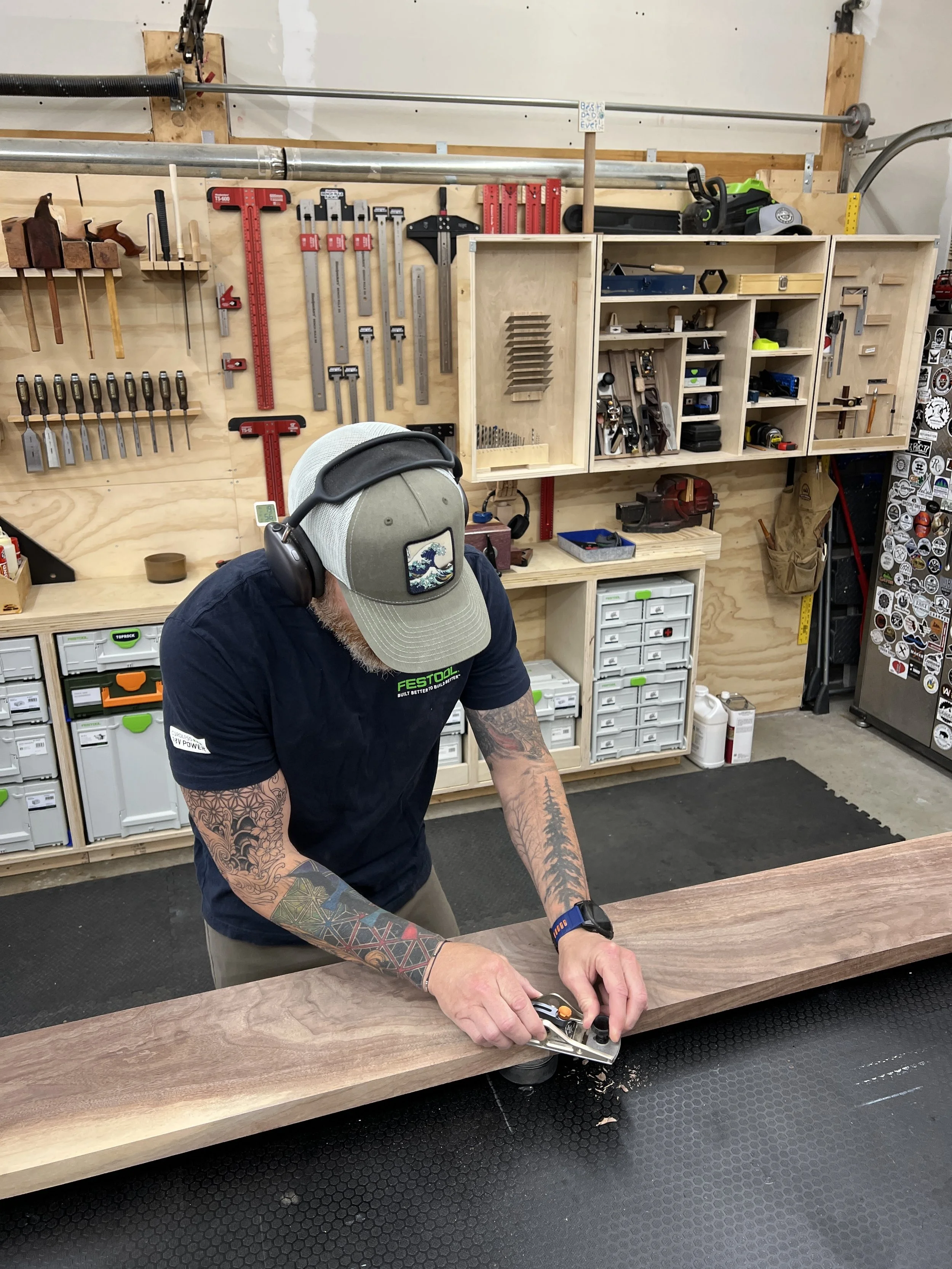A man working in a woodworking workshop, using a hand plane on a wooden board. The workshop has tools hanging on a pegboard and storage bins in the background.