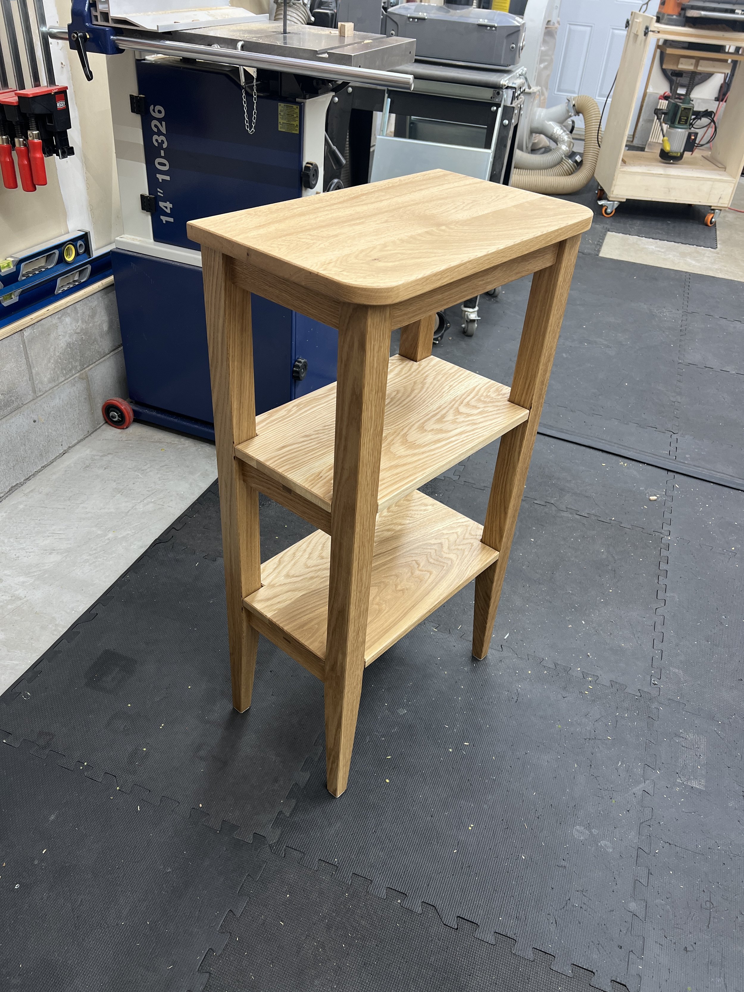 A three-tier wooden cart with a flat top, middle shelf, and bottom shelf, situated on a black rubber mat in a workshop. In the background, there are various woodworking tools and equipment.