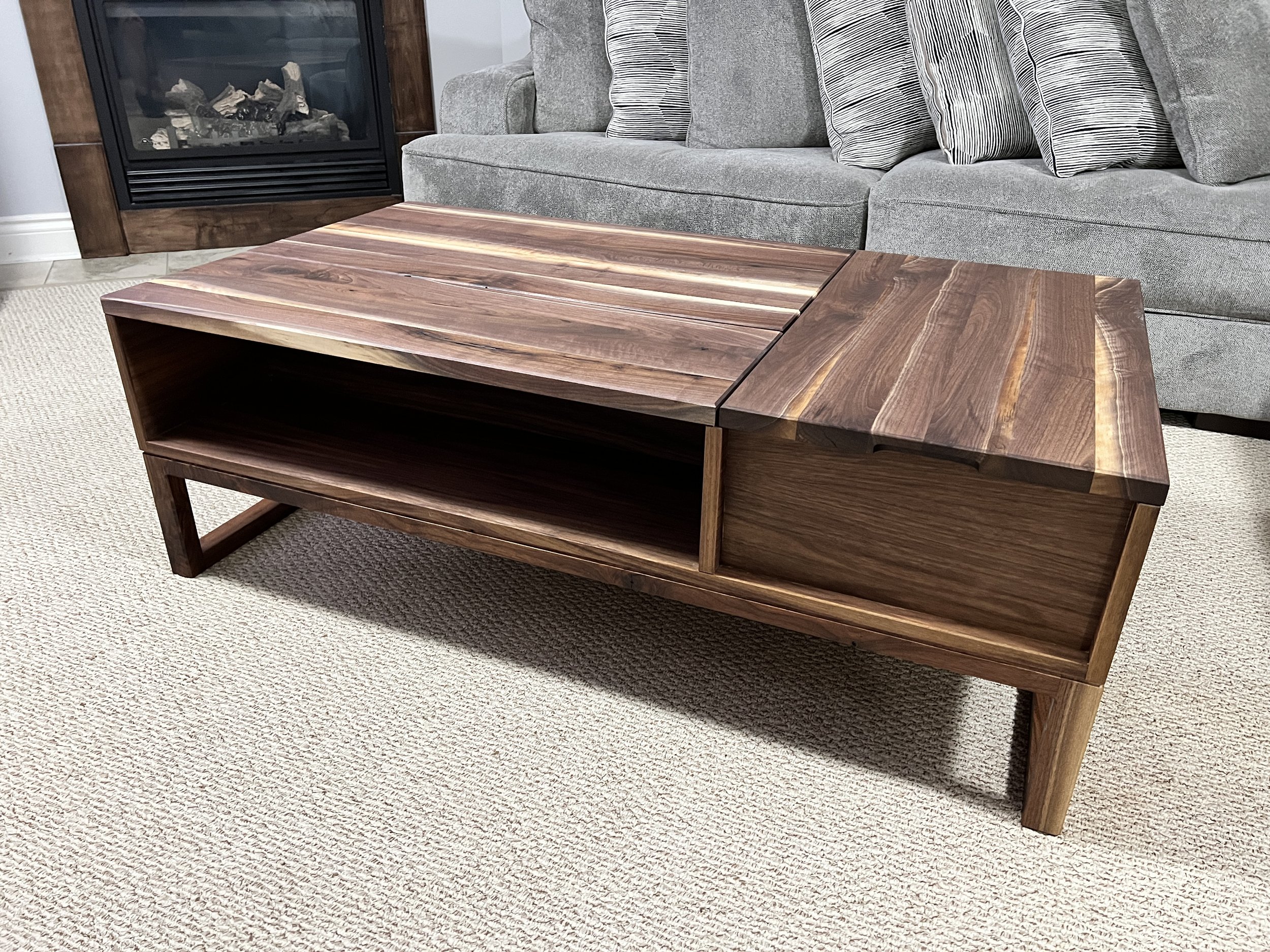 A wooden coffee table with a slatted top in front of a gray fabric sofa with striped pillows, in a room with a fireplace to the left and a beige carpeted floor.