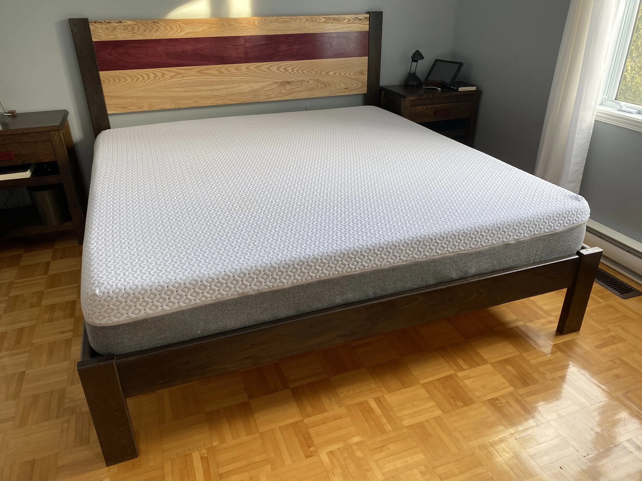 A hotel bedroom featuring a bed with a white textured mattress, a wooden headboard with natural and red stains, and dark wood side tables. One side table has a reading lamp, a tablet, and a closed book. The room has light green walls, a window with w