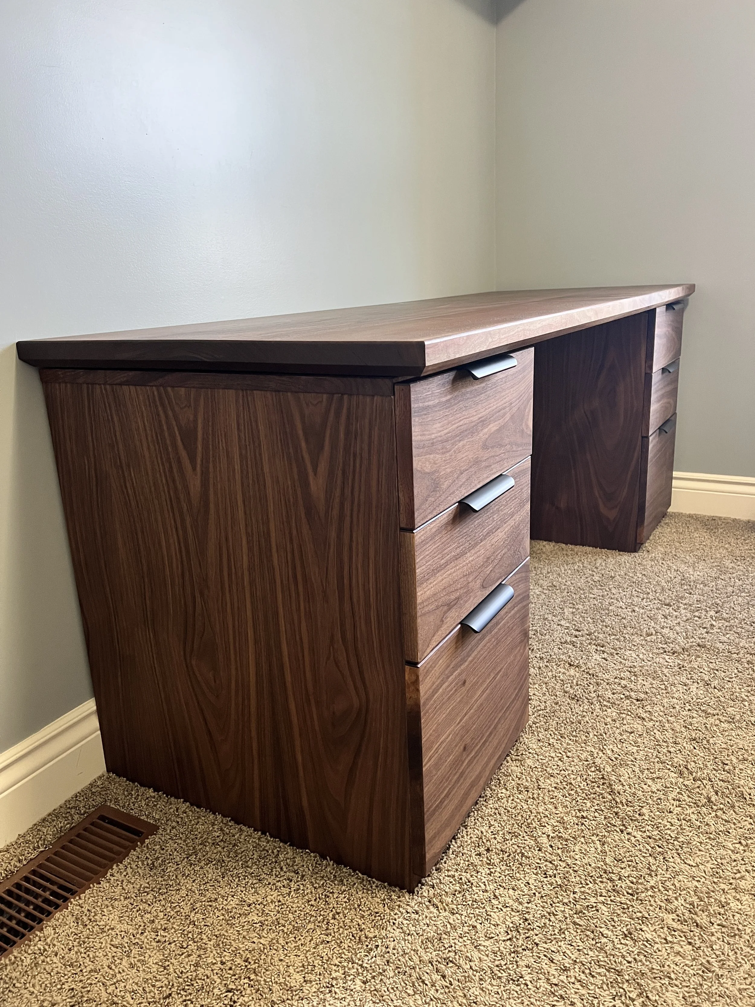 A wooden dresser with multiple drawers and silver handles, placed against a light-colored wall in a room with beige carpet.
