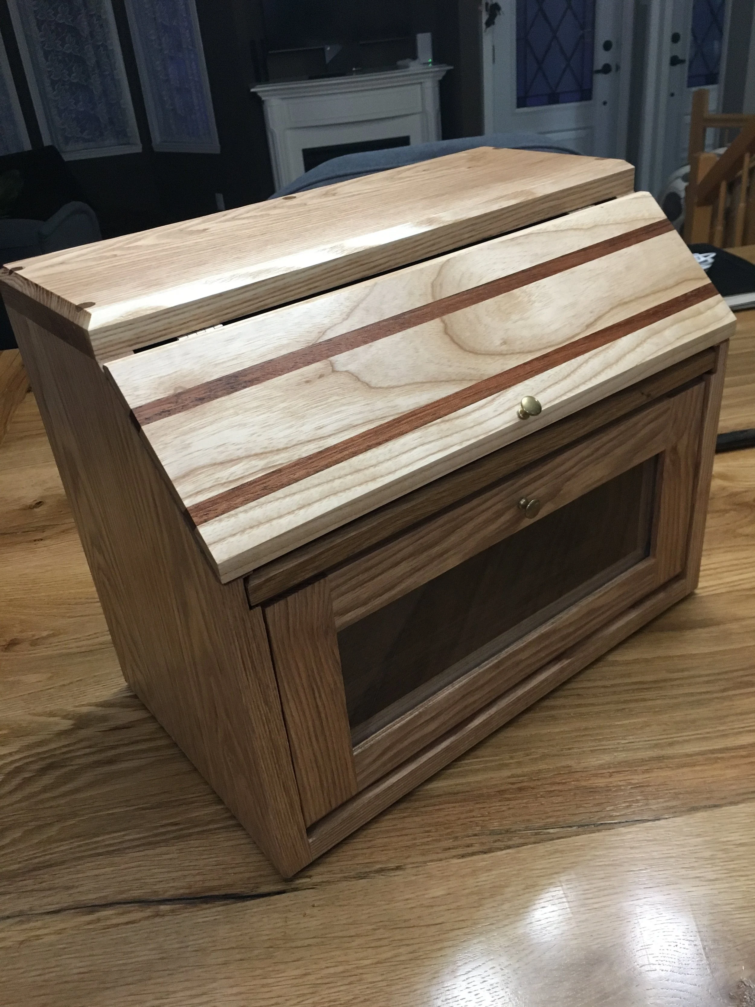 A wooden box with a lid, featuring a striped pattern with natural wood tones, sitting on a wooden surface in a room with a fireplace and furniture in the background.