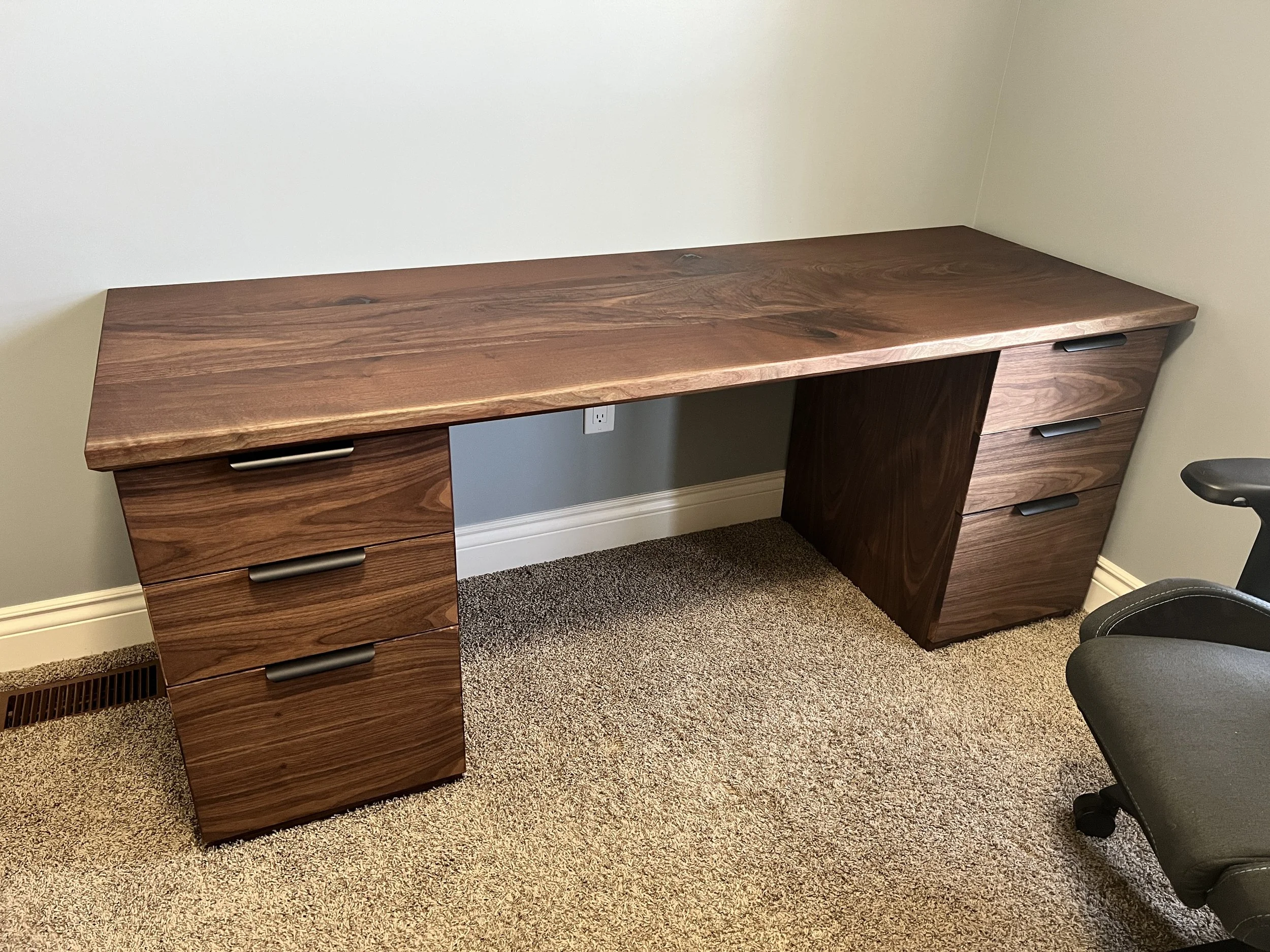 A wooden desk with a smooth top and multiple drawers, situated in front of a beige wall and a carpeted floor, with part of a black office chair visible on the right.