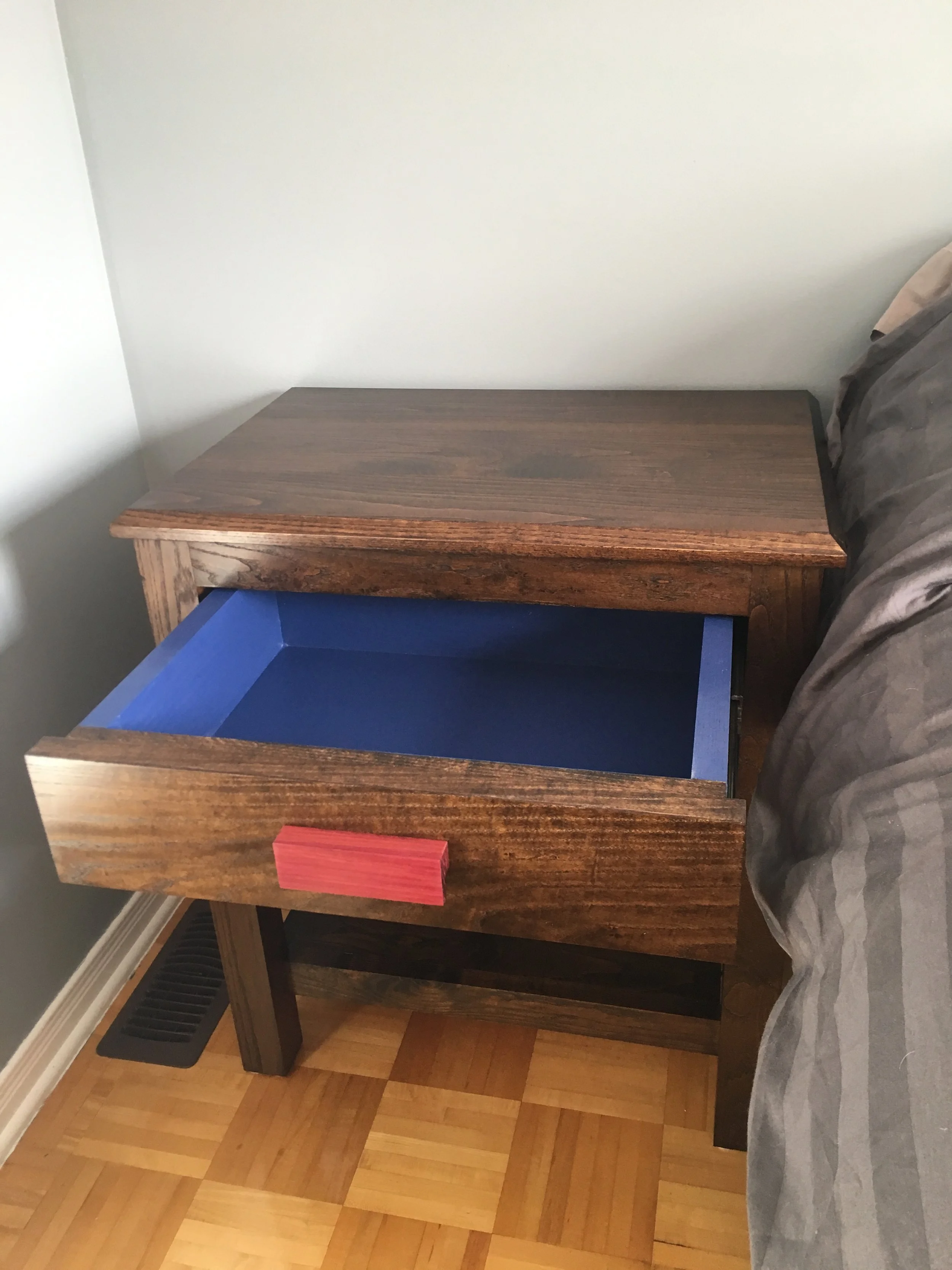 A wooden nightstand with a blue drawer and a small red object on top, located beside a bed with gray bedding, in a room with hardwood flooring and a white wall.
