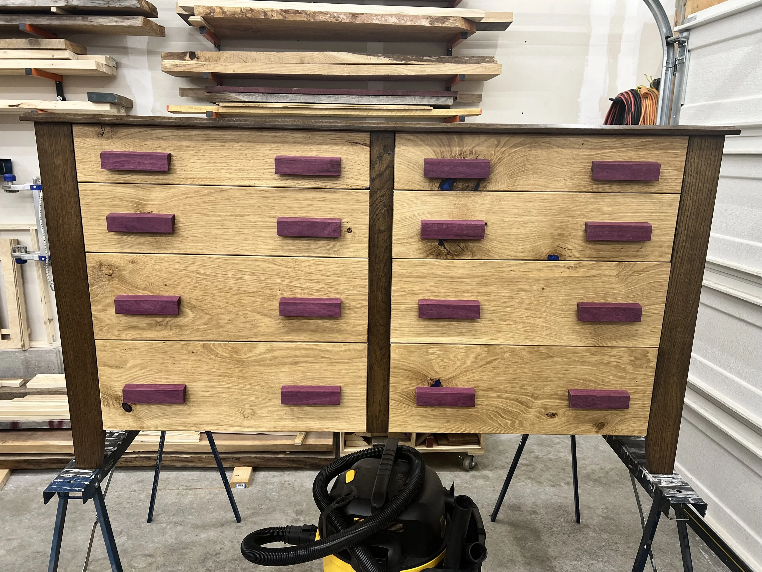 A wooden dresser with purple drawer handles, in a woodworking workshop with sawhorses, shelves with wood planks, and a vacuum cleaner on the floor.