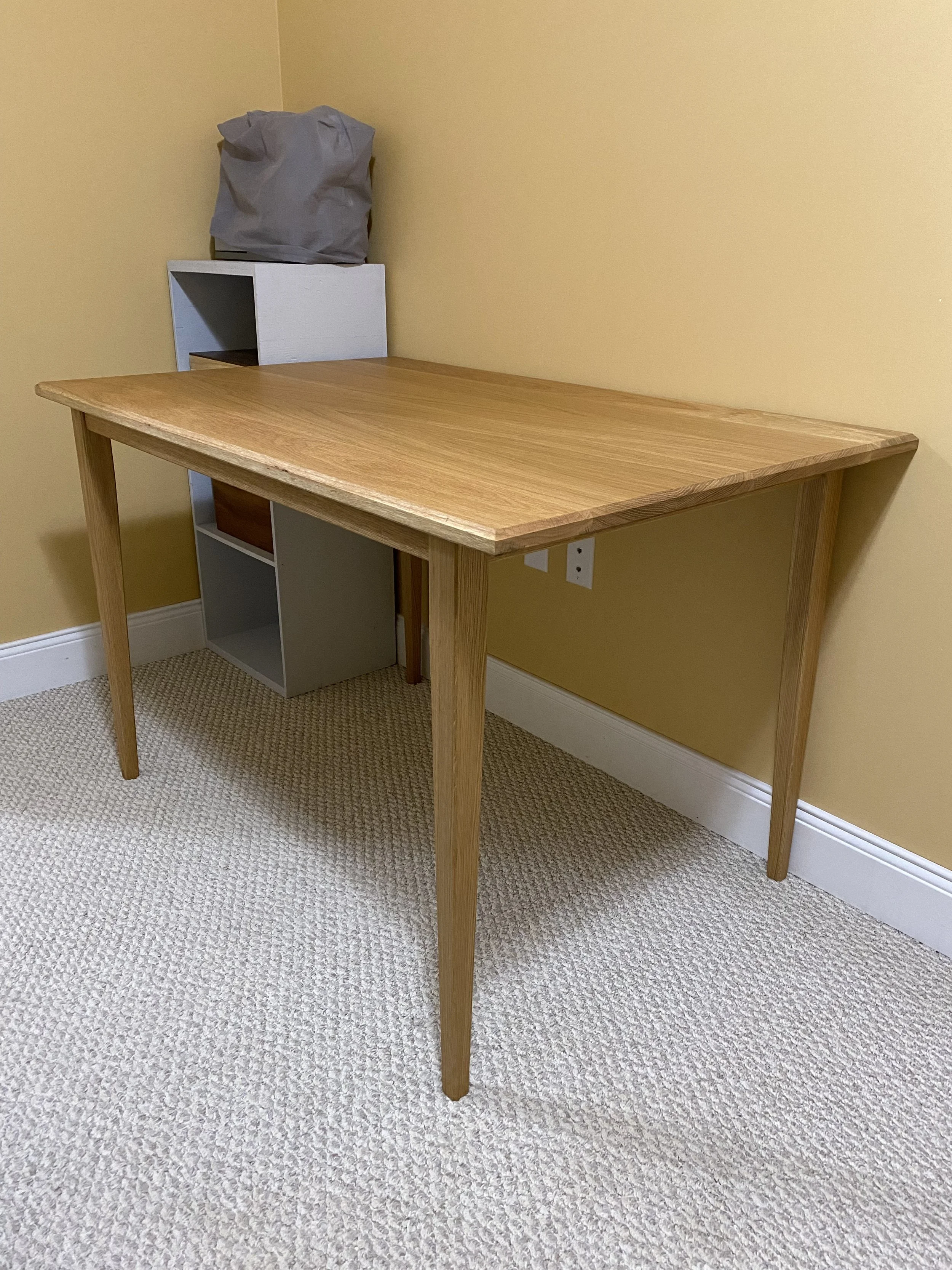 A wooden desk with four legs positioned against a yellow wall, with a small white and gray shelving unit on top, and a gray bag placed on the shelf.