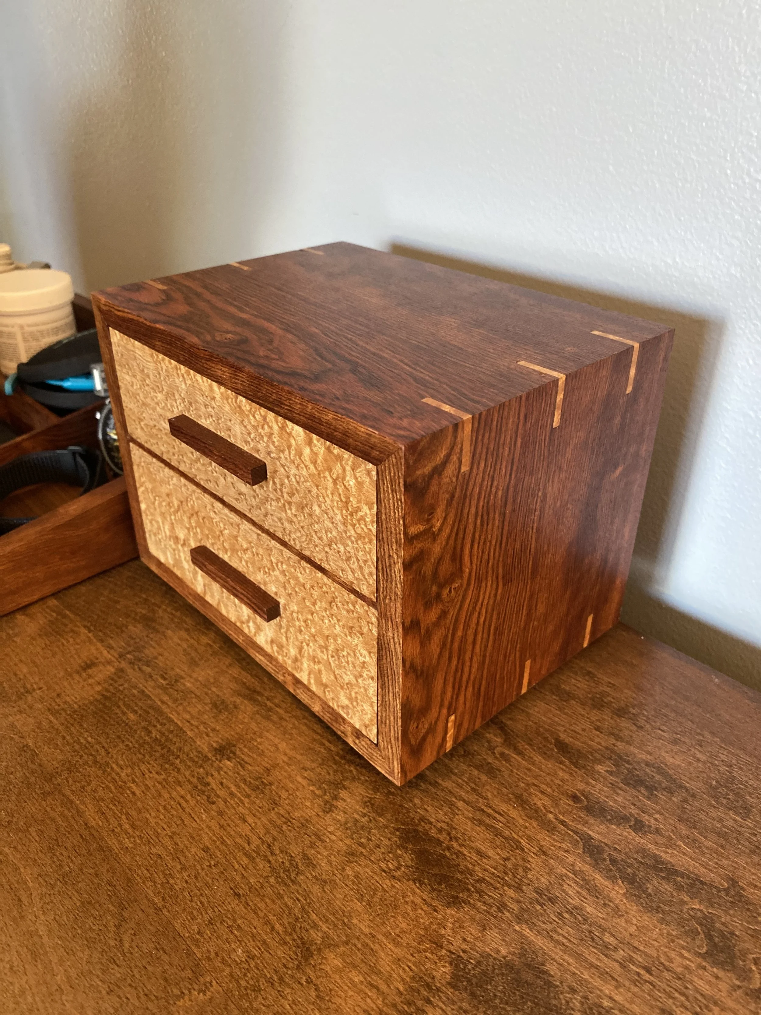 A wooden bedside table with two drawers, made of a lighter and darker wood, placed on a wooden surface in front of a light-colored wall.