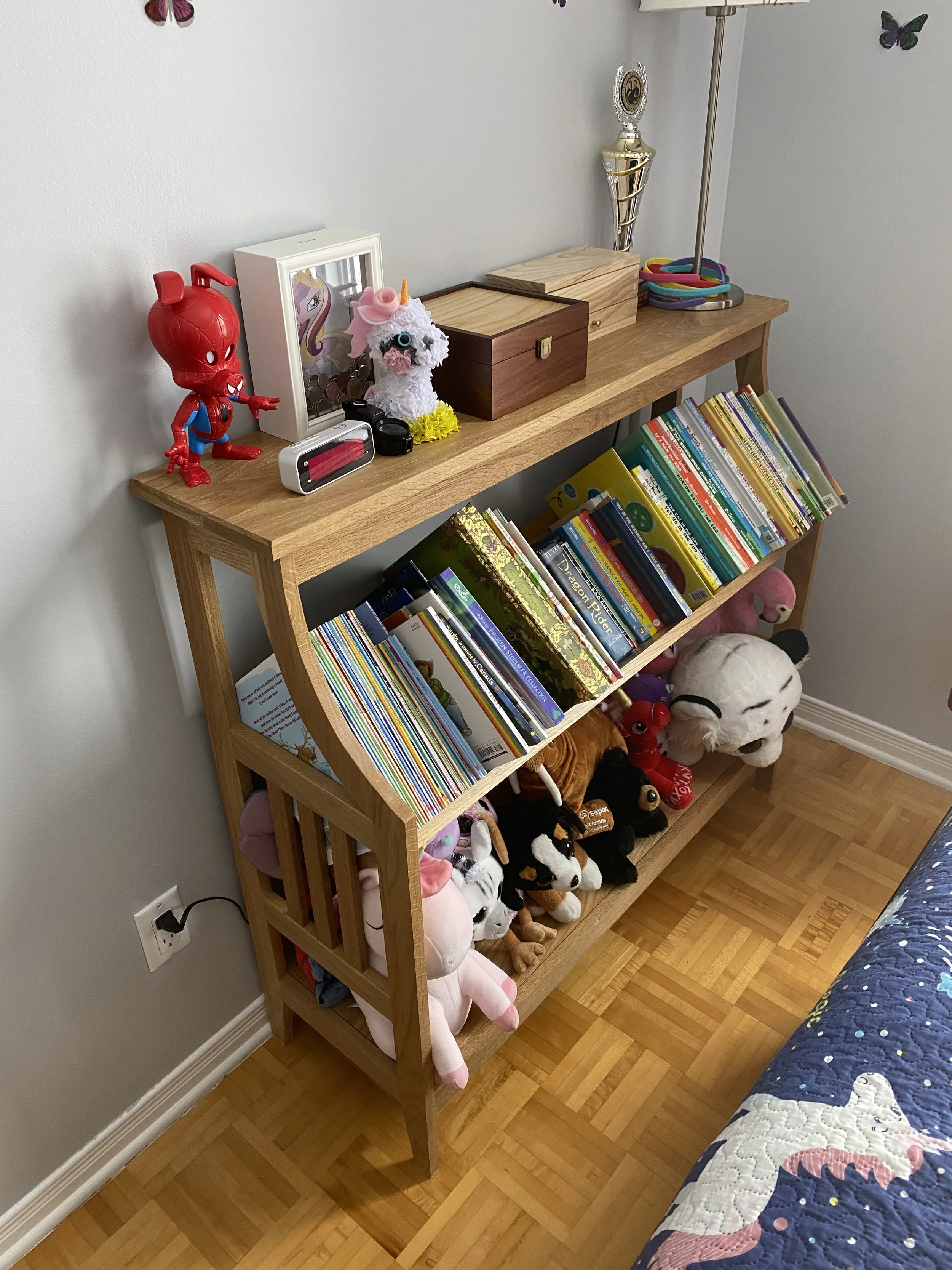 A wooden bookshelf filled with children's books on the middle shelf, plush stuffed animals on the bottom shelf including pandas, a pig, queen of hearts, and other characters, a small collection of colorful bangles in a dish, a lamp, and trophies and 