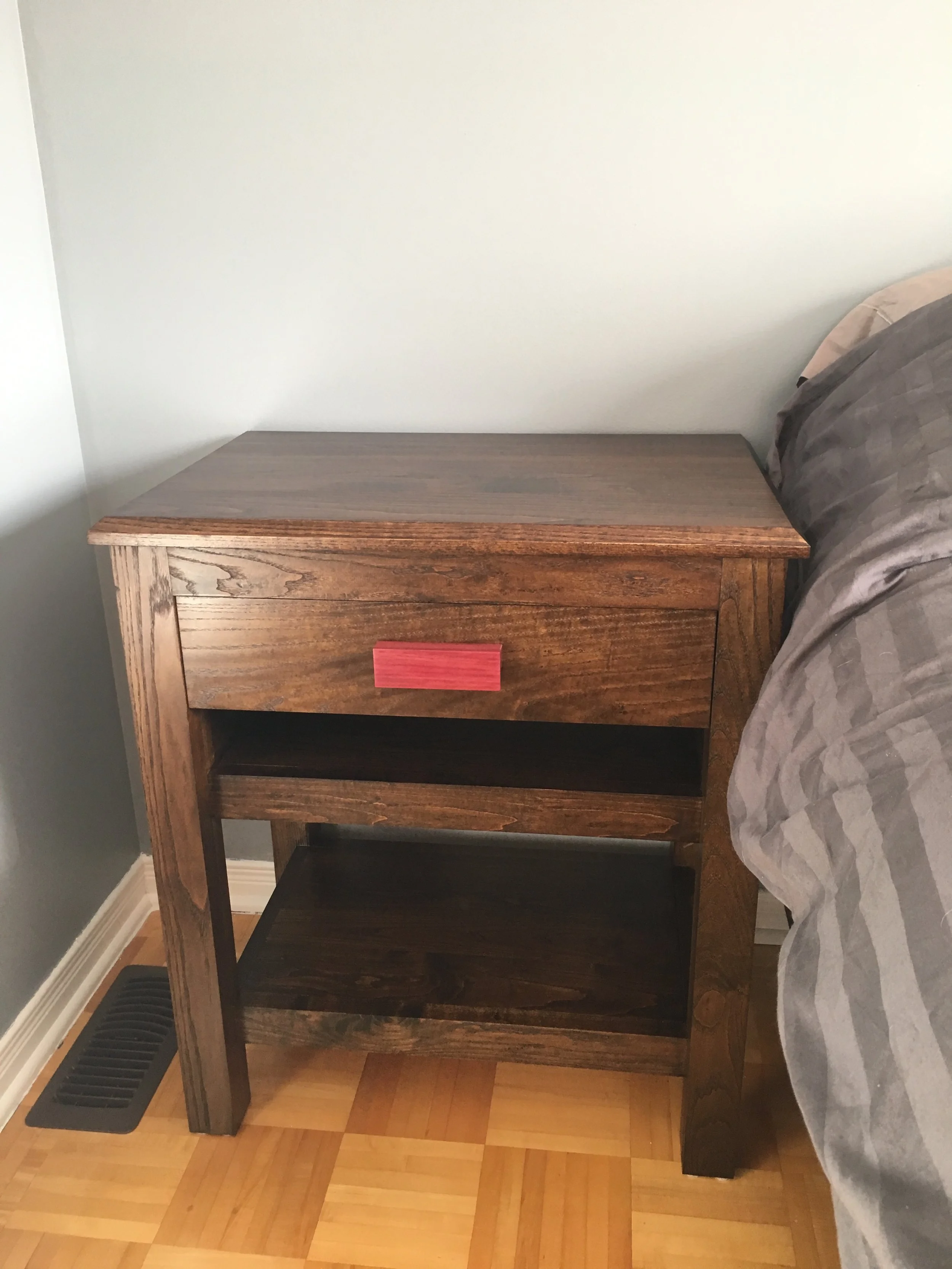 A wooden nightstand with an open shelf at the bottom and a drawer with a red handle, placed next to a bed with a gray quilted blanket. The nightstand is against a wall with a light gray color, nearby a vent on the floor, and a hardwood floor.