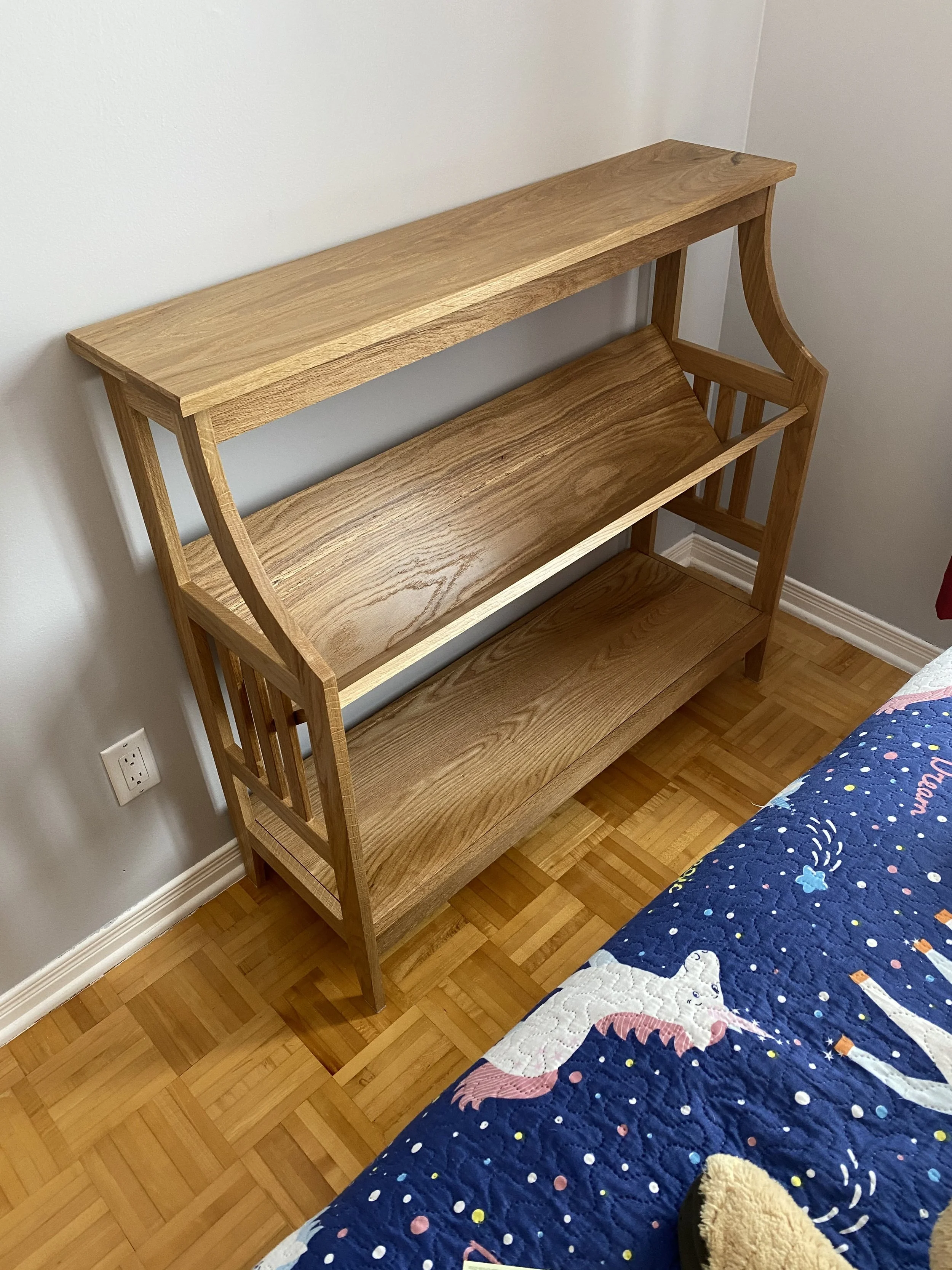 Wooden bookshelf with three shelves next to a bed with a colorful quilt, on a wooden floor and white wall.