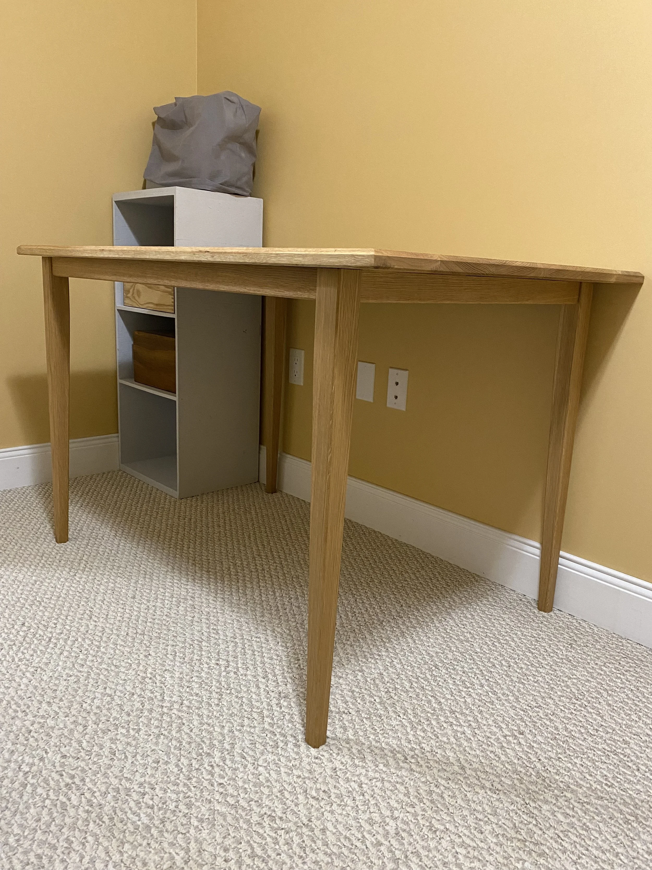 A wooden desk with a light wood finish, partially pushed against a yellow wall in a room with beige carpet. There is a small gray cubby storage unit underneath the desk, topped with a gray bag on top of a white box.