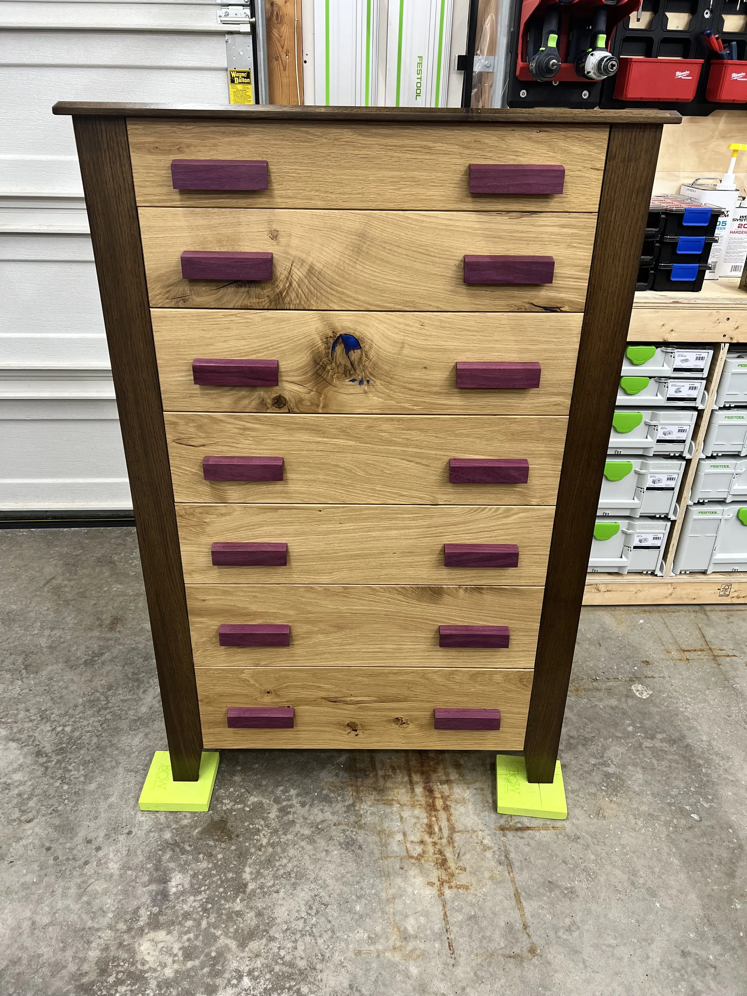 A wooden dresser with dark brown sides and purple drawer handles, standing on concrete floor with yellow painter's tape underneath each leg.