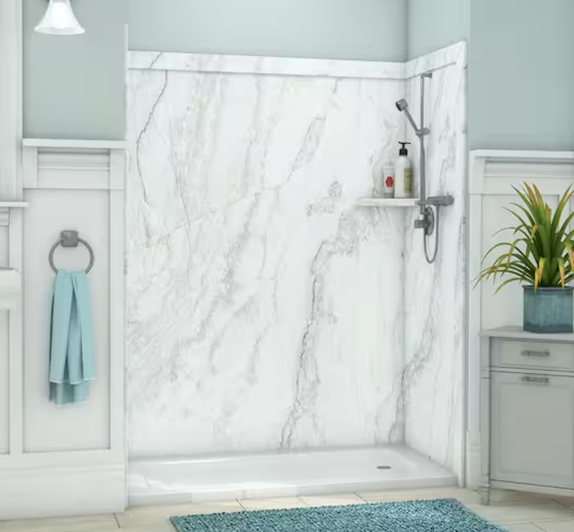 A bathroom shower area with white marble walls, a showerhead, and a small shelf holding shampoo and body wash bottles. A light blue towel hangs on a towel ring next to the shower, and a potted plant is placed on a gray cabinet nearby.