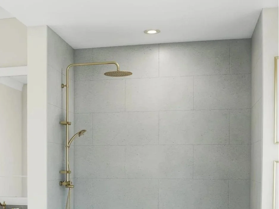 A modern, minimal shower area with a ceiling-mounted rain shower head and a wall-mounted handheld shower head, both in a gold finish, against large, light gray tiles.