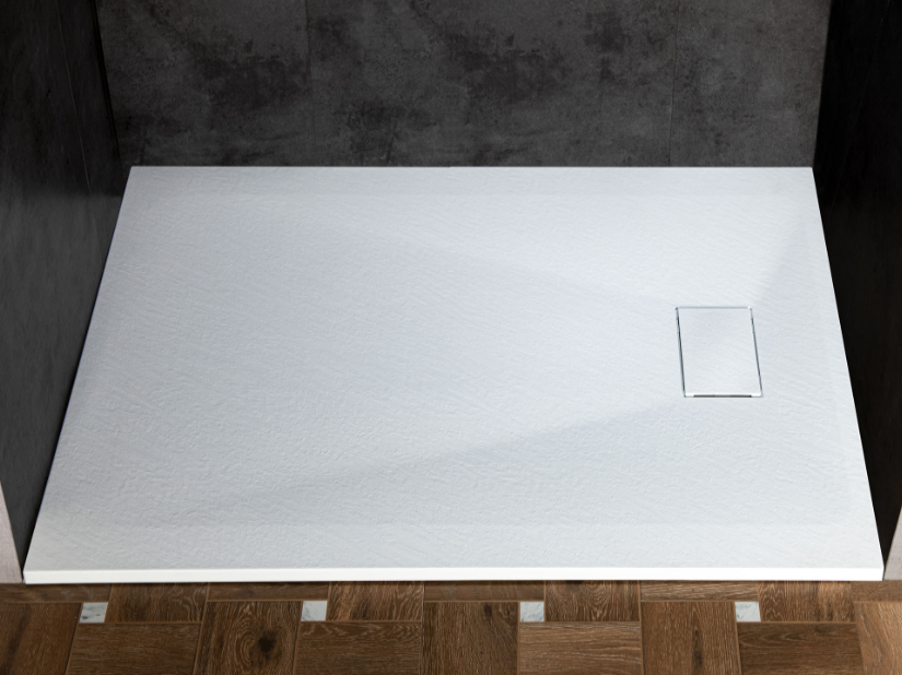 Empty white shower base with a small access panel, set against dark wall and wooden flooring.