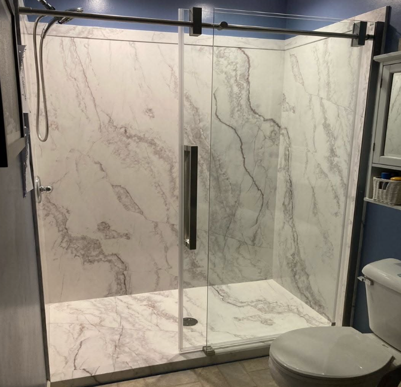 A bathroom shower with marble walls and floor, a glass door with a metal handle, a wall-mounted shower head, and a toilet to the right.