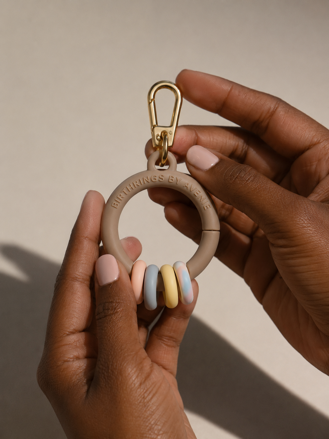 Person holding a beige keychain with colorful silicone rings and a gold carabiner clip, with the inscription 'BIRTHINGS BY AVENUE' on the ring.