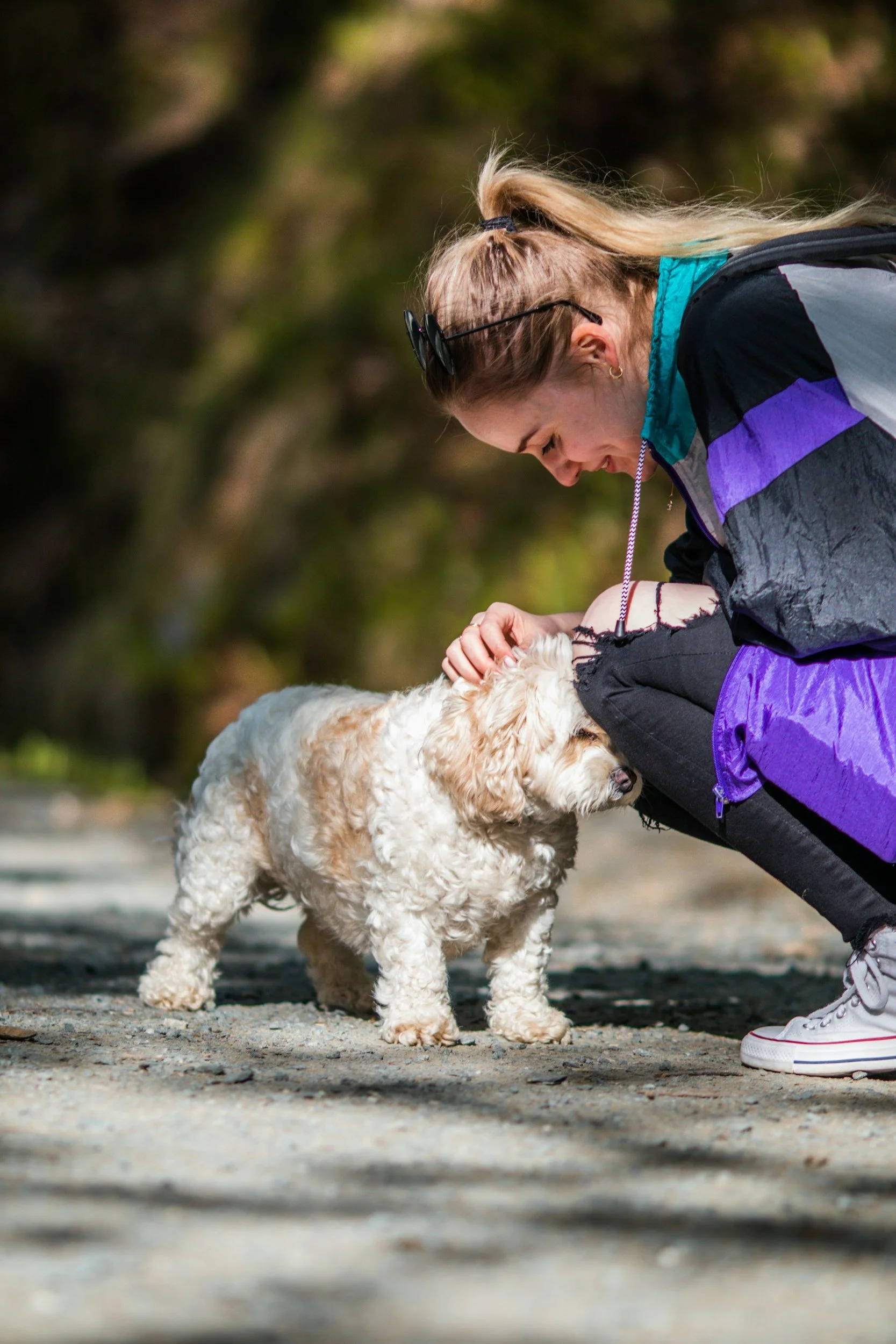A woman with blonde hair tied in a ponytail, wearing sunglasses on her head, a colorful jacket, and sneakers, bending down to interact with a small, fluffy, light-colored dog on a dirt or gravel path outdoors.