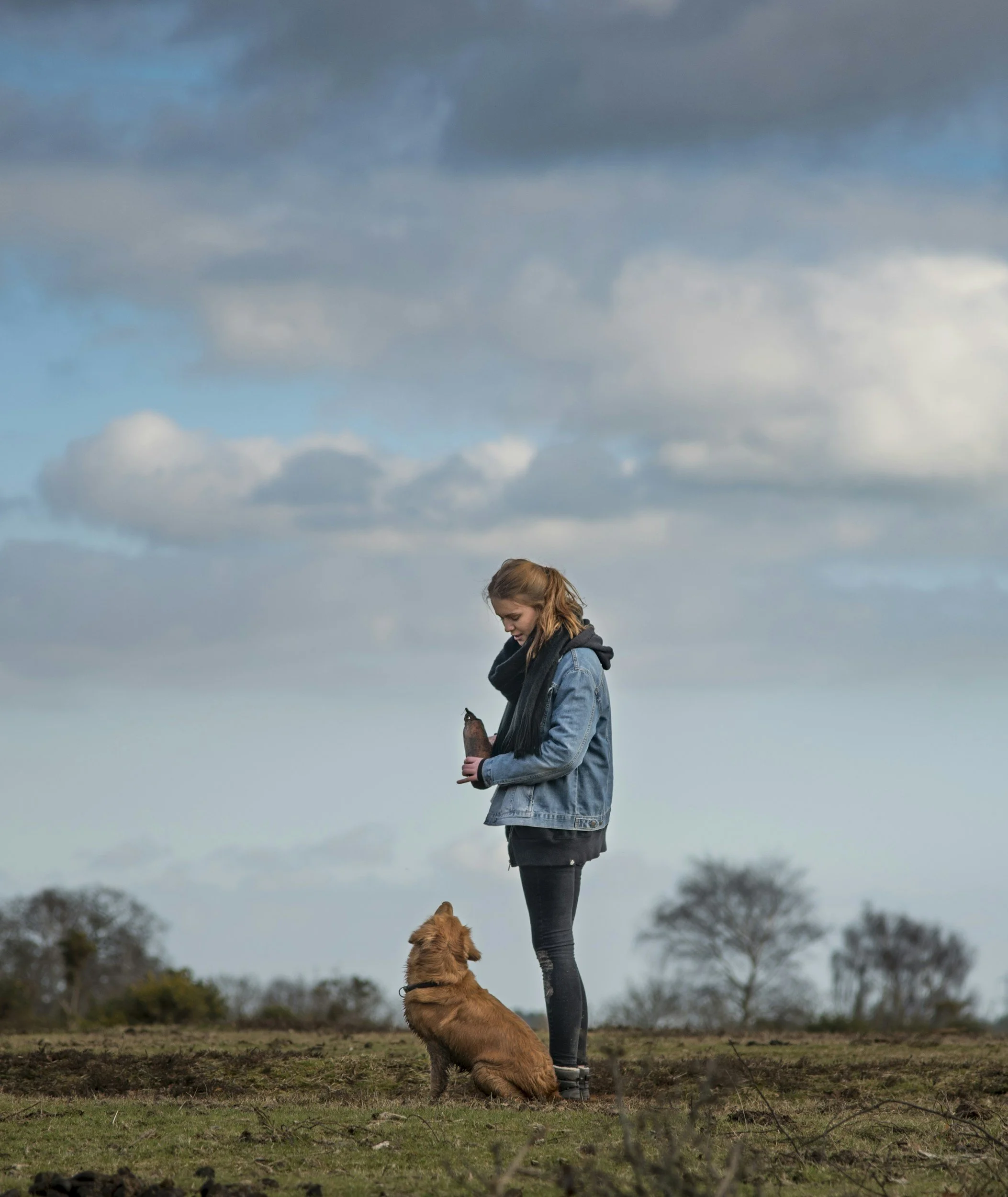 A young woman standing outdoors in a field, looking at her phone, with a golden retriever sitting at her feet. The sky is cloudy with patches of blue.