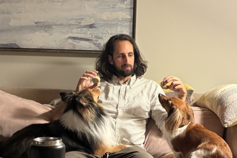 A man with shoulder-length dark hair and a beard is sitting on a sofa, holding two cookies, one in each hand. He is surrounded by two dogs, one on each side of him, looking at him. The room has a large, abstract painting on the wall behind him and a blanket or pillow on the sofa.