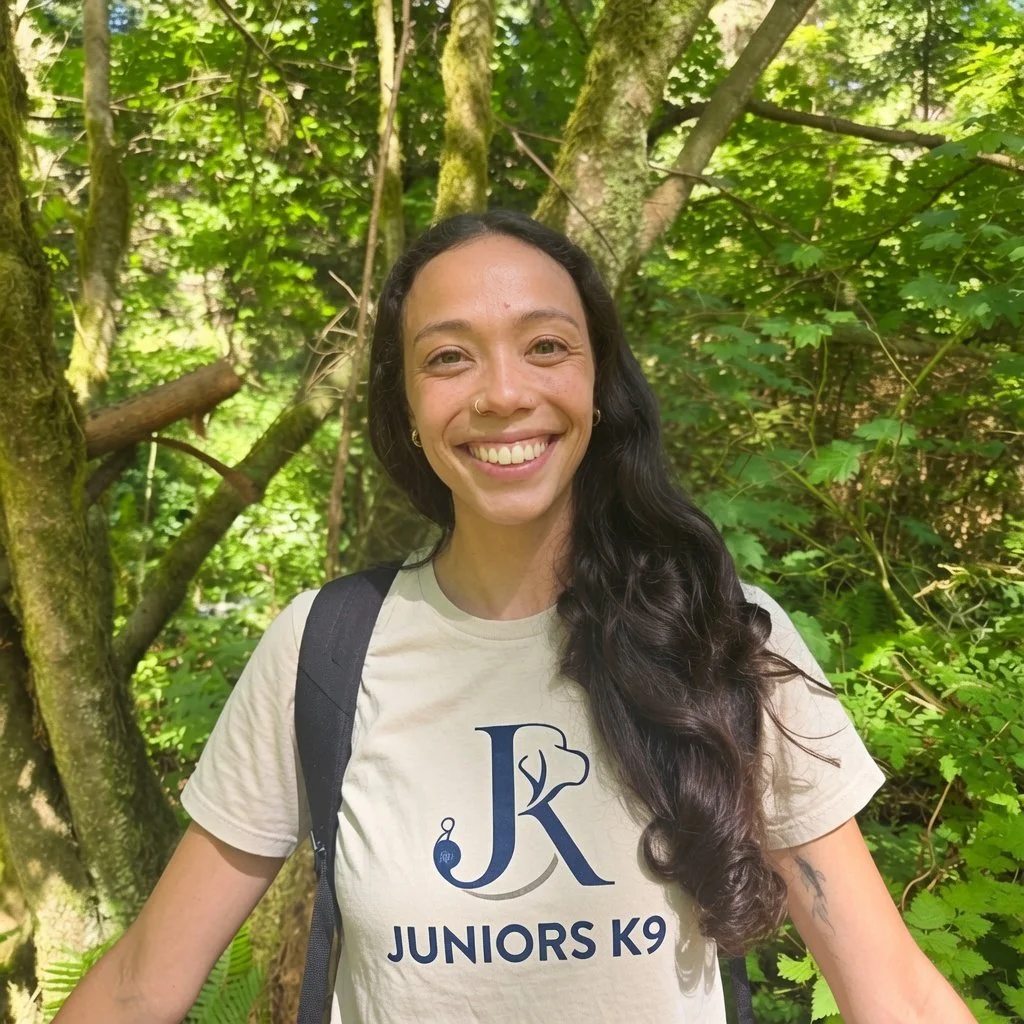 A smiling woman with long dark hair, wearing a beige t-shirt with 'JUNIORS K9' printed on it, standing outdoors in a lush green forest with trees and foliage in the background.