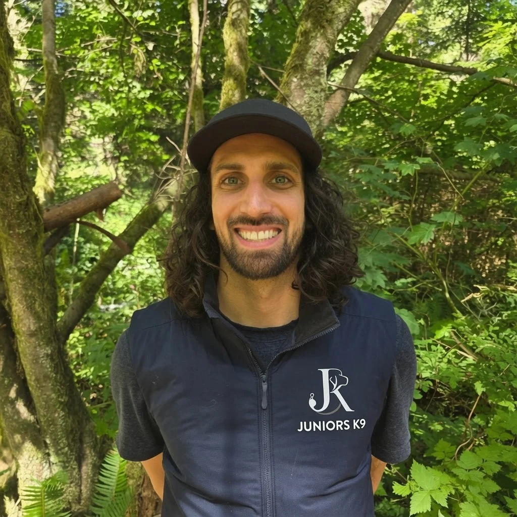 A man with curly dark hair, a beard, and light skin smiling outdoors in a lush green forest, wearing a black cap and a black vest with a Juniors K9 logo.