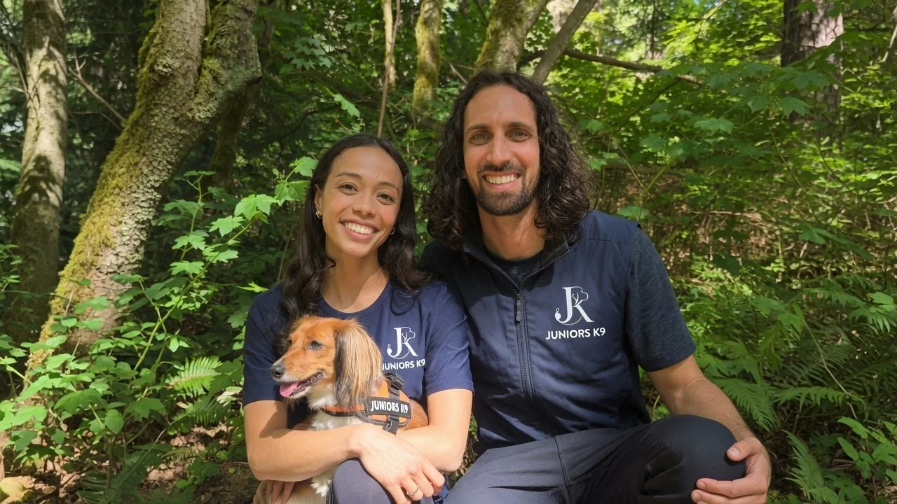 A man and woman sitting in a forest with green foliage, smiling at the camera, while the woman holds a small dog wearing a harness.