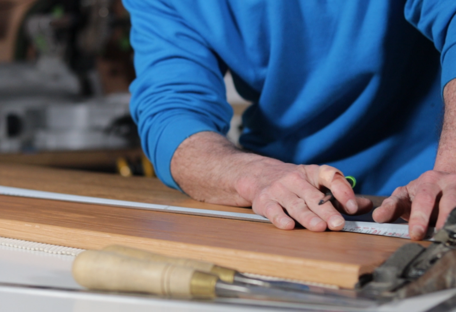 A person wearing a blue shirt measuring a piece of wood on a workbench with a metal ruler, a pencil, and a hammer nearby.