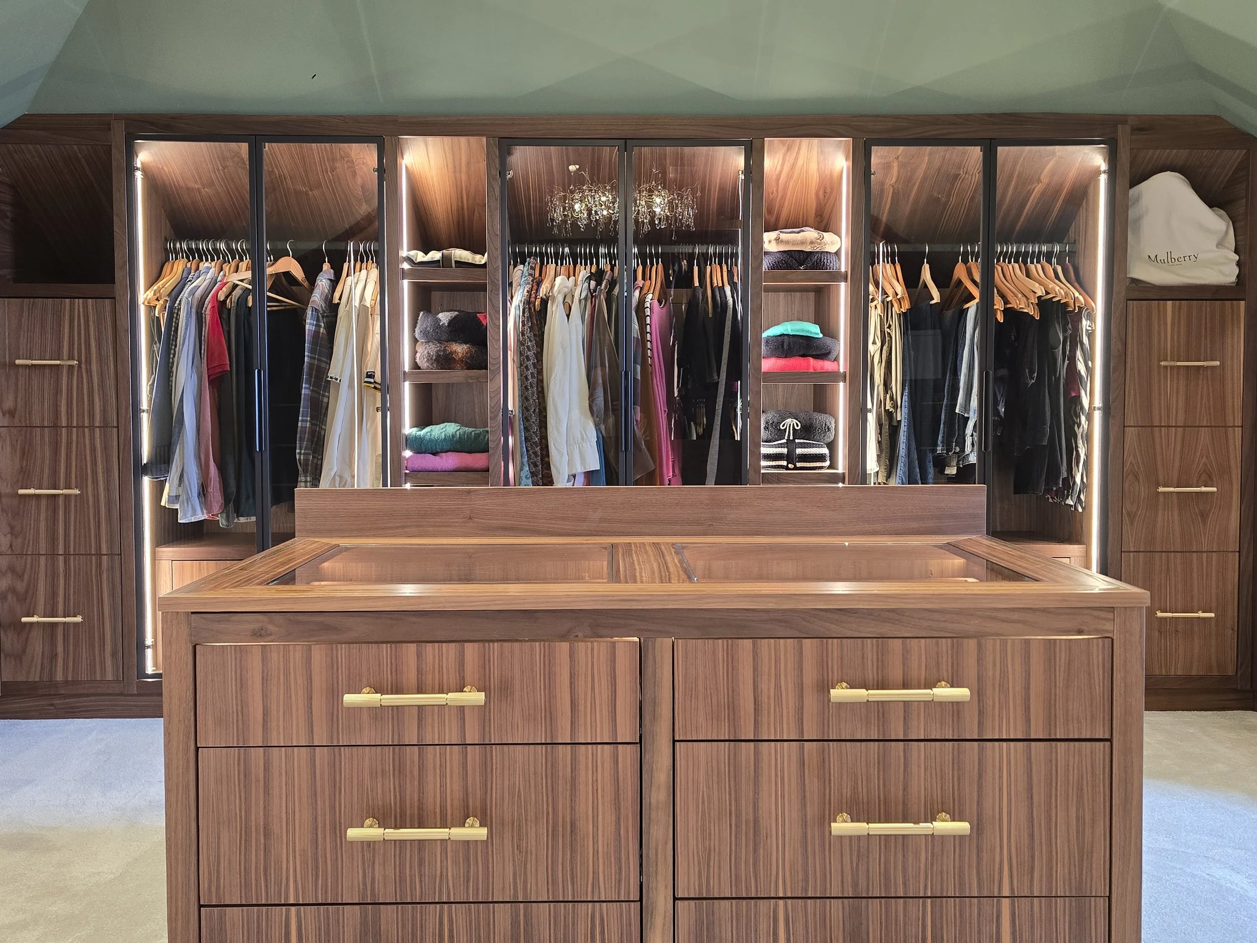 A large wooden wardrobe with glass doors, containing hanging clothes, folded sweaters, and a jewelry chandelier inside. There are drawers with gold handles at the bottom and a Mulberry bag on the side shelf.
