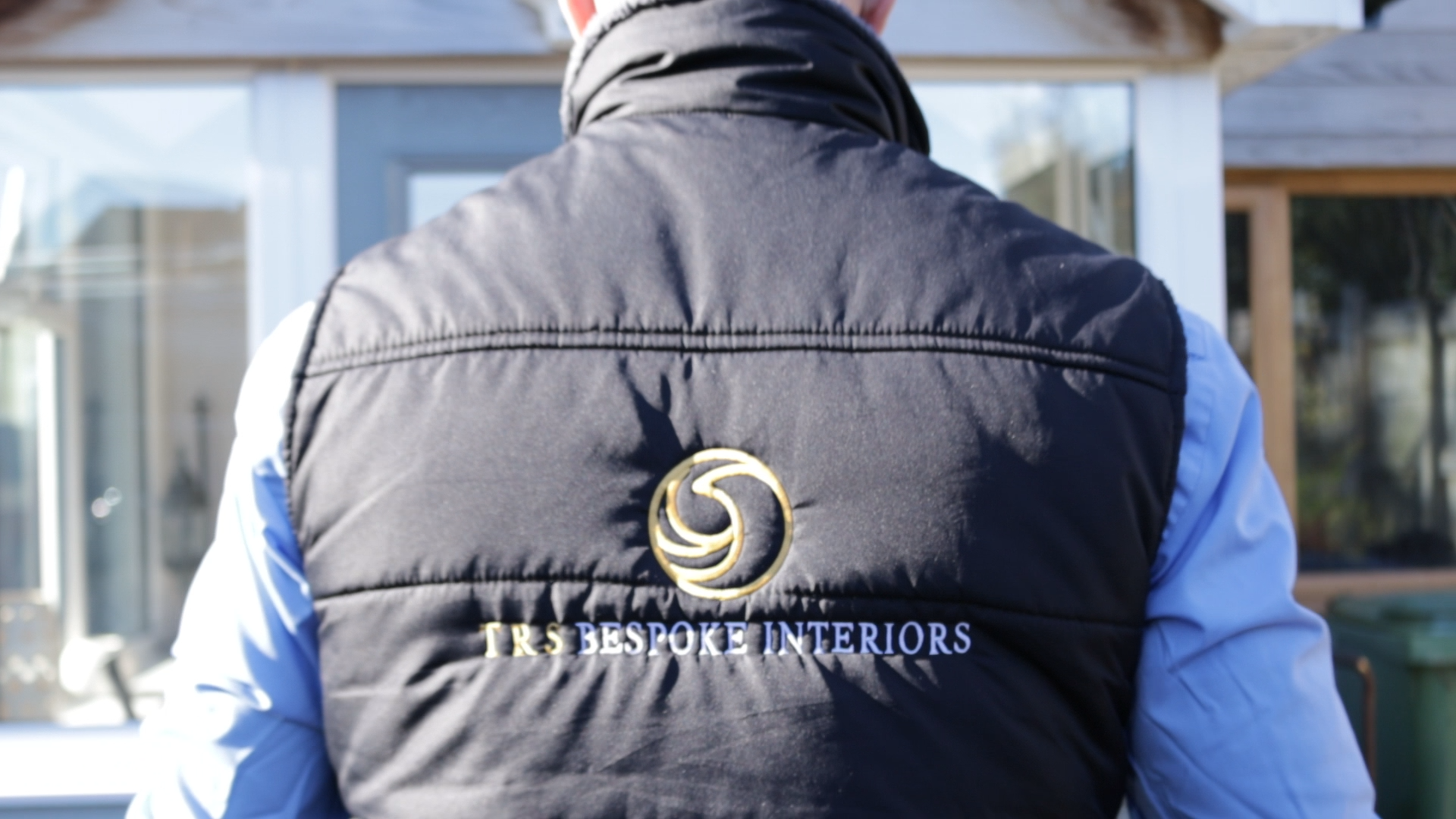Back of a person wearing a black vest with embroidered gold and white text and logo, standing outdoors near a building with glass windows.