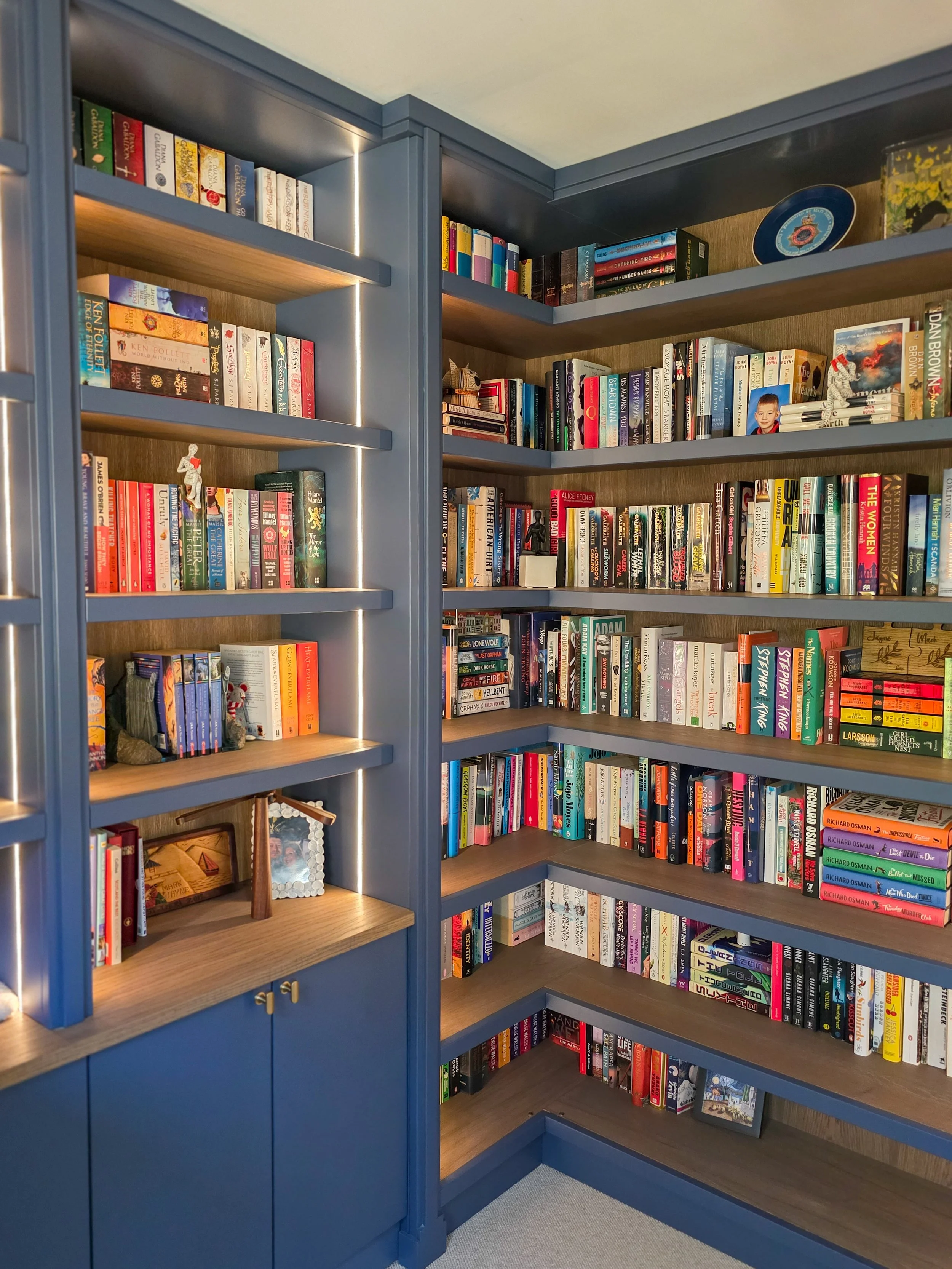 Blue wooden bookshelf filled with books, with some shelves decorated with small figurines and a framed photograph, in a cozy room.