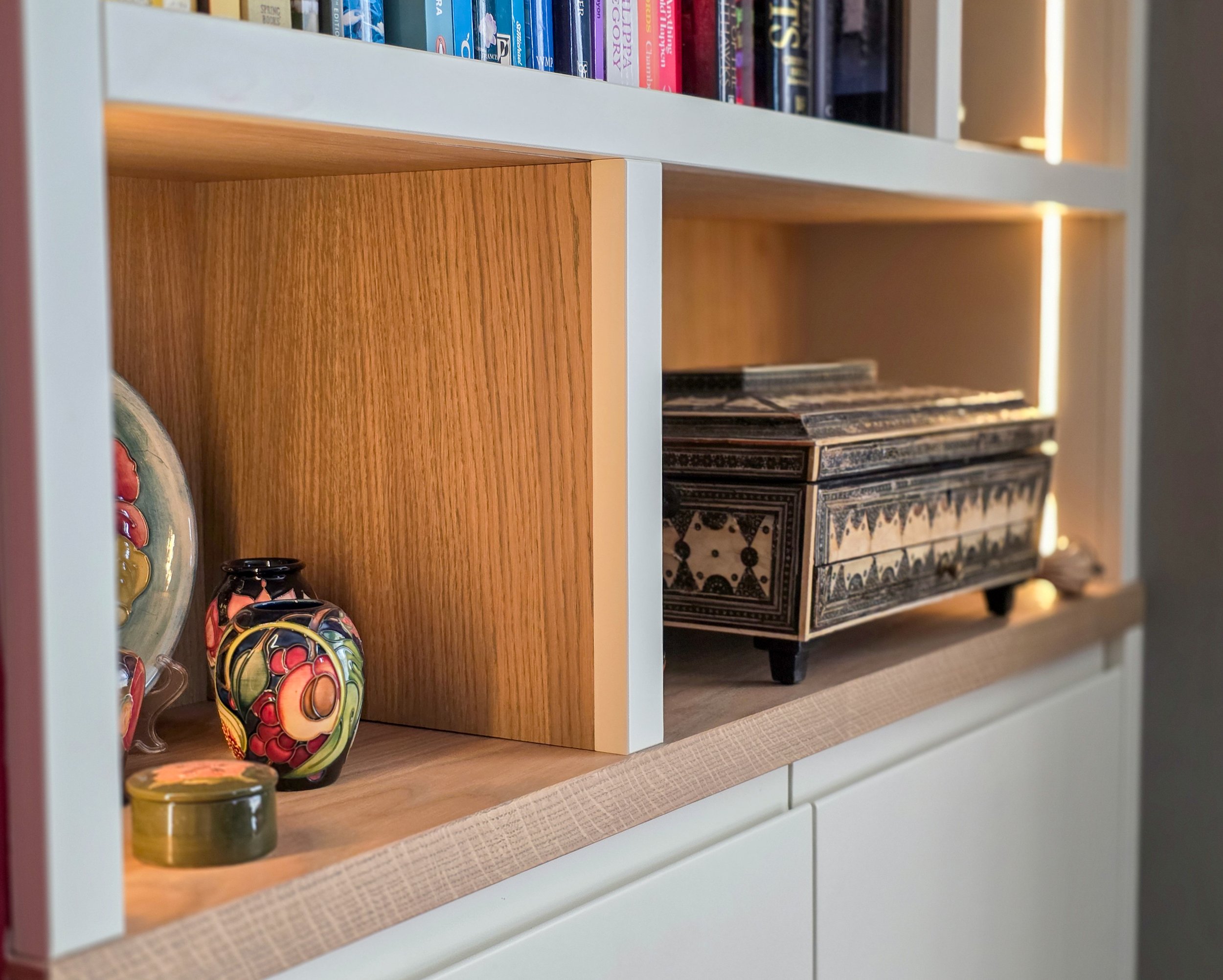 Close-up of a bookshelf with a white frame, wooden middle section, and decorated with colorful ceramic skulls and small containers in the lower shelf, with books on the upper shelves.