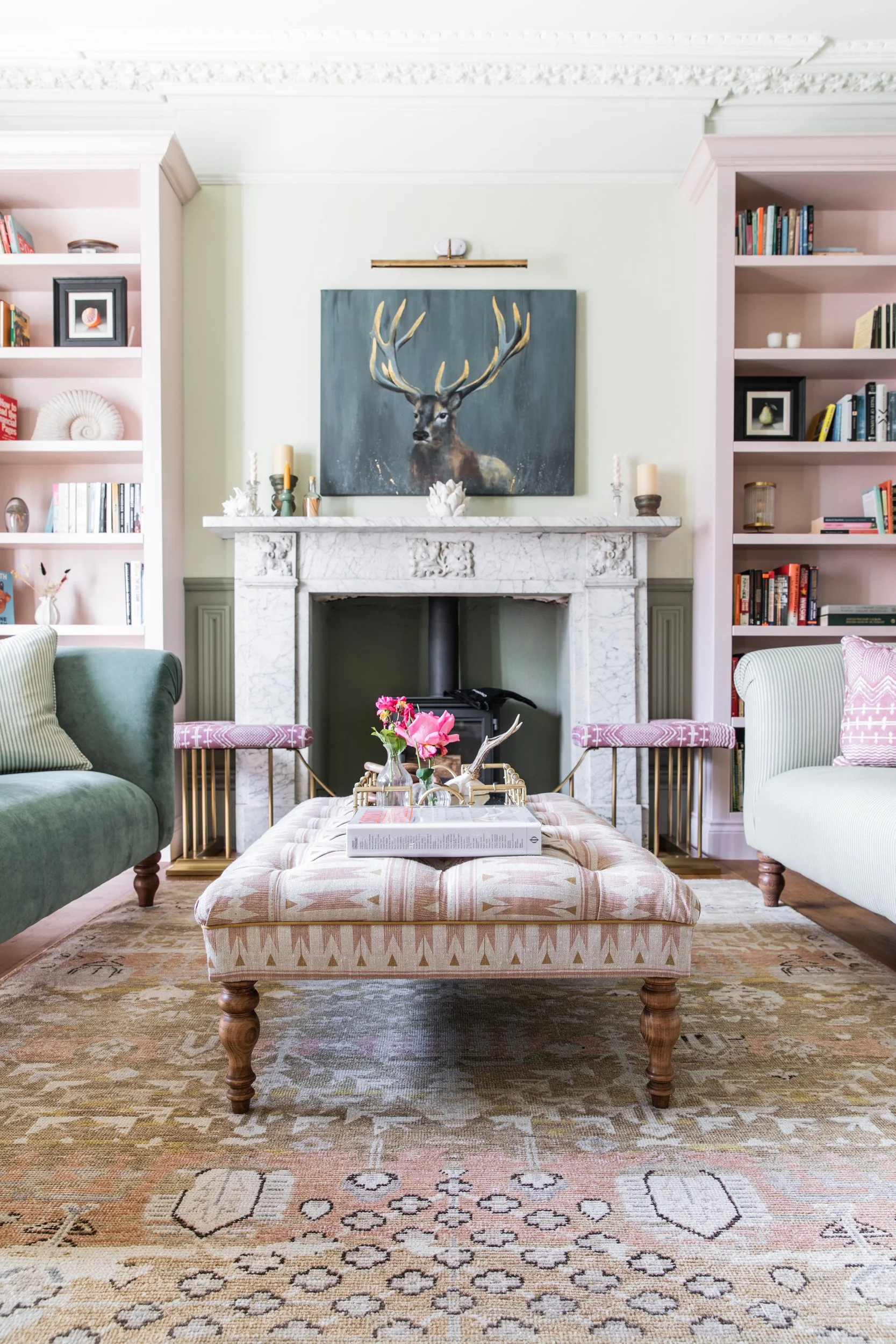 A living room with a white marble fireplace, a painting of a deer with large antlers above it, and built-in shelves filled with books and decorative items. There are chairs, a pink upholstered ottoman, and a carpet on the floor.