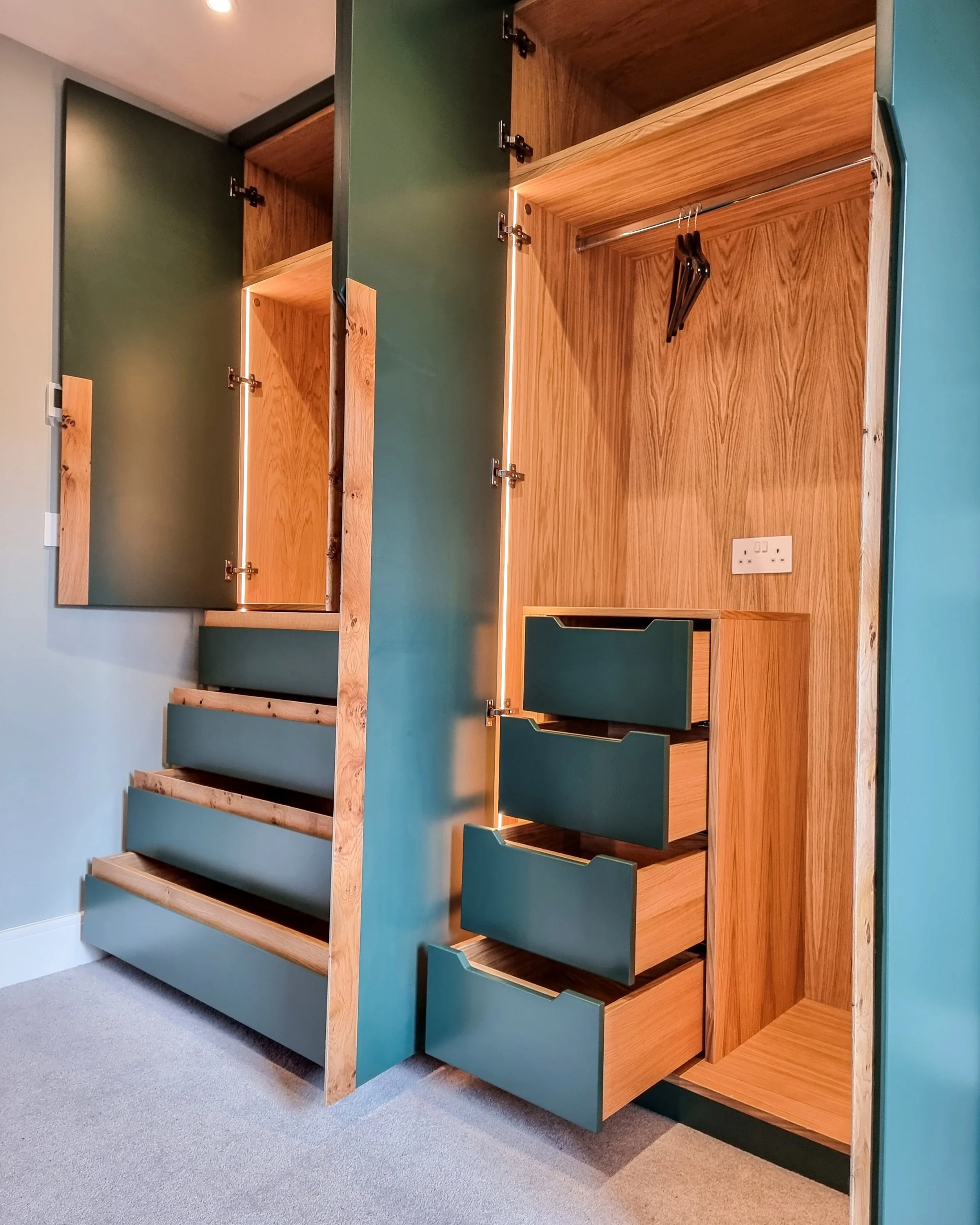 Open wooden closet with teal doors, metal hanging rod, and three wooden hangers inside. The closet has a partial view of an electrical outlet on the back wall.