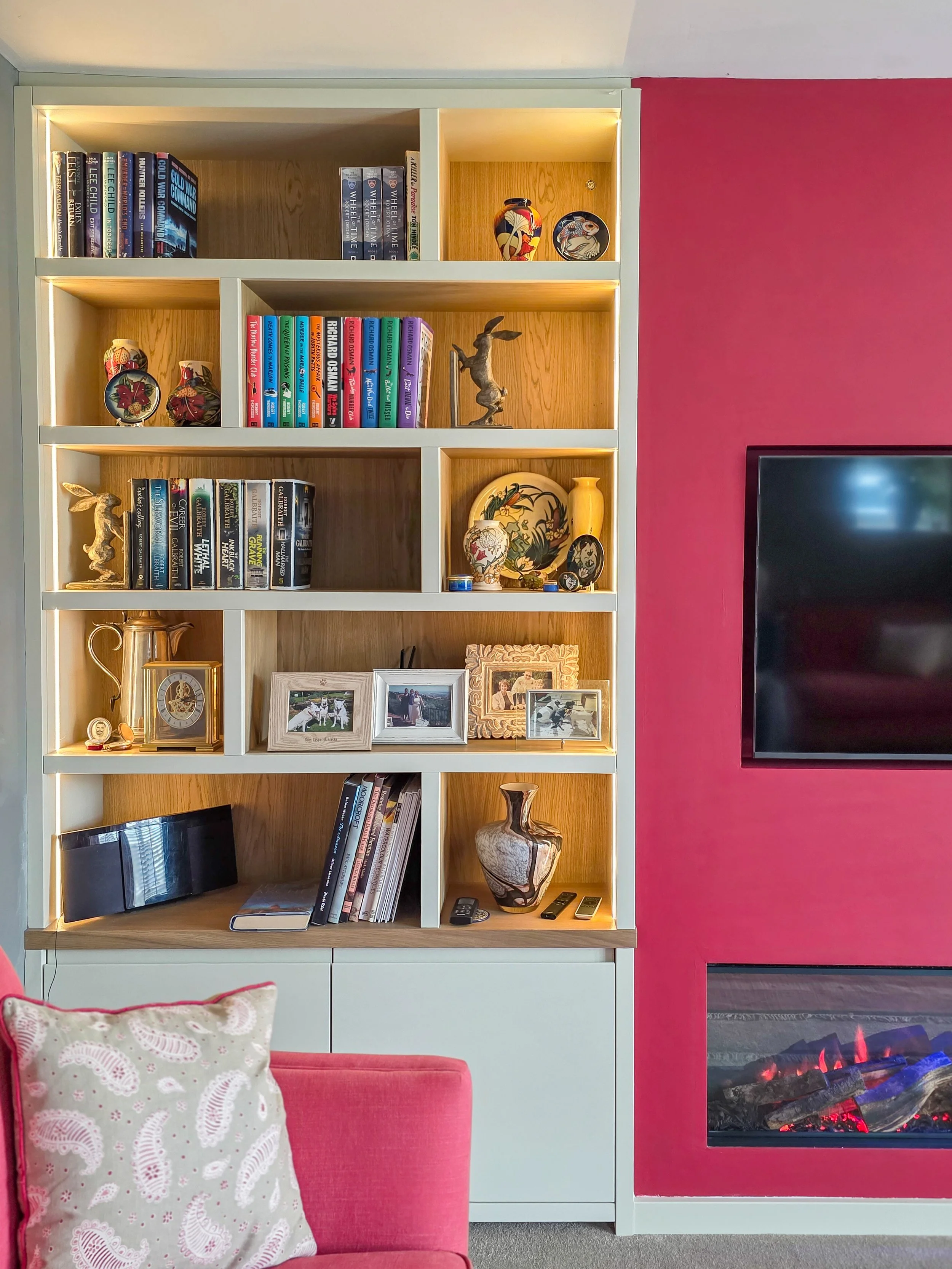 A modern living room with a large white bookshelf filled with books, decorative items, and framed photos. To the right, there is an electric fireplace with realistic flames, set into a bright pink wall. A pink couch with a white decorative pillow sits in the foreground.