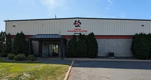Exterior of a building with a sign and tall bushes in front, parking lot and grassy area nearby.