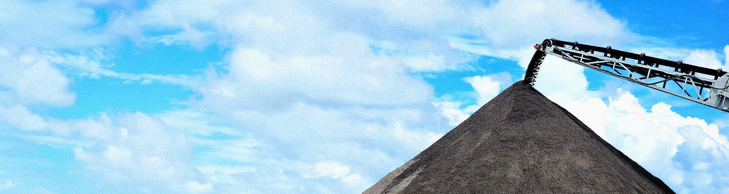 A large conveyor belt is depositing black gravel or crushed stone onto a tall mound under a partly cloudy sky.