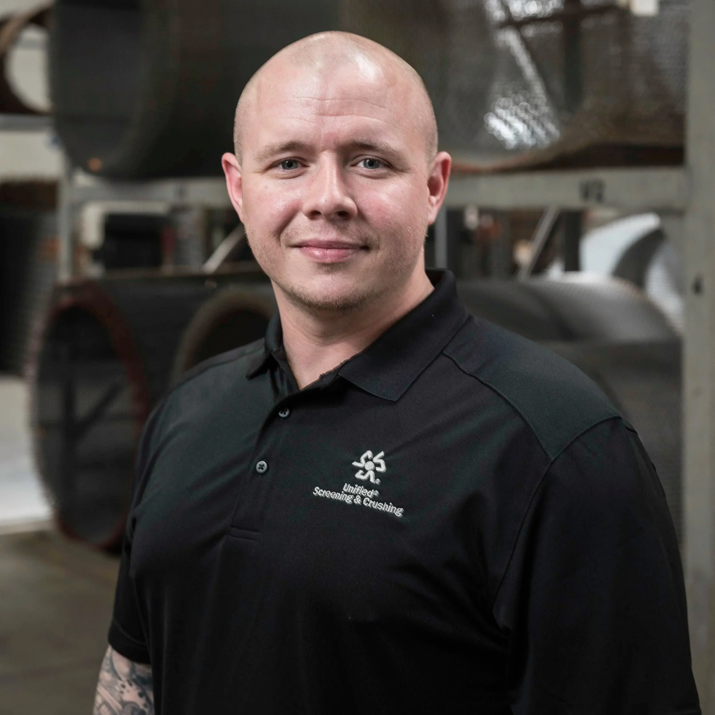 A man with a shaved head and blue eyes wearing a black shirt with white text and logo, standing in a setting with blurred industrial background.