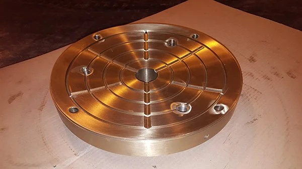 A circular metallic component with a central hole and multiple smaller holes arranged in a pattern, placed on a wooden surface.