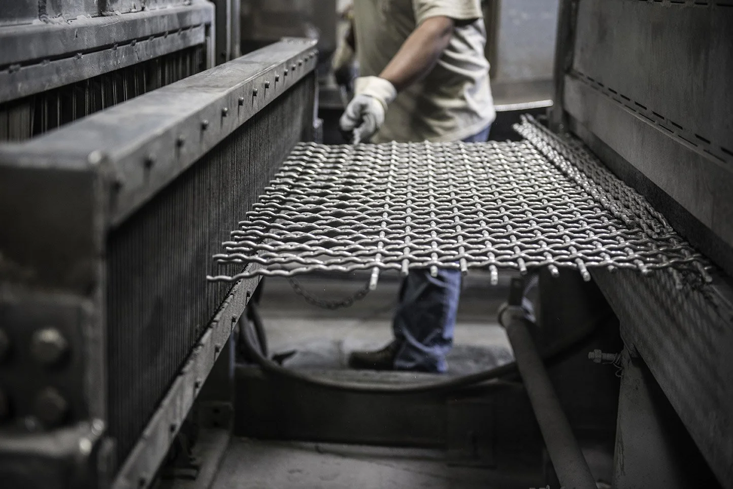 A worker in gloves handling hot metal objects on a conveyor in an industrial foundry or manufacturing facility.