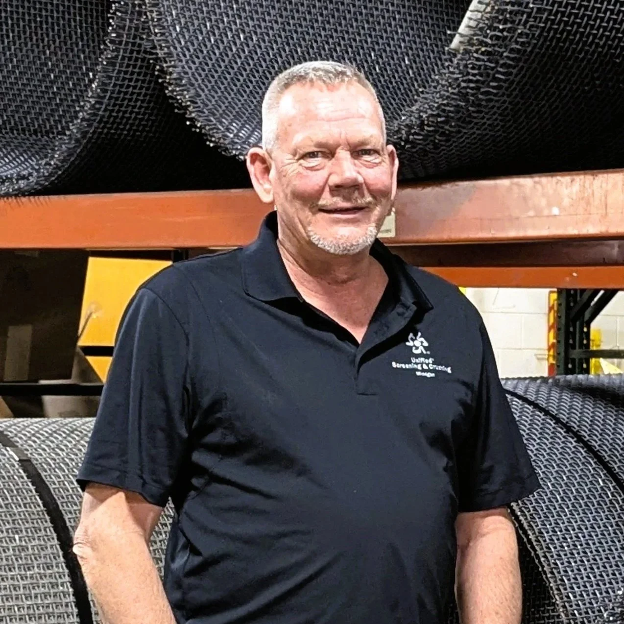 A middle-aged man with short gray hair and a beard, smiling, wearing a black polo shirt with a logo on the chest, standing in front of industrial metal mesh and storage shelves in a warehouse setting.