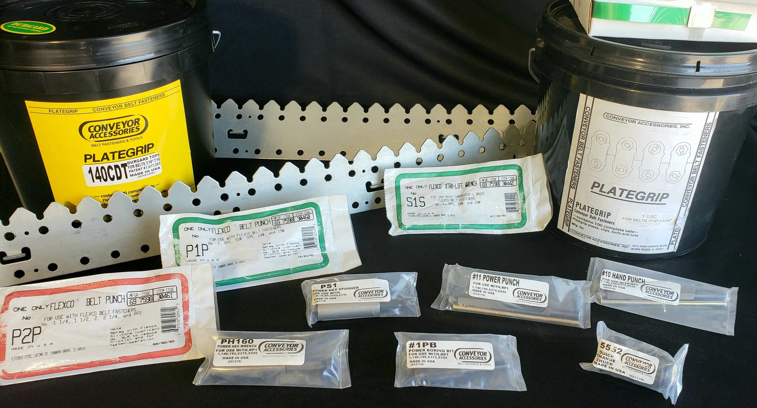 Various packages of conveyor belt fasteners and tools on a black surface, including black plastic buckets with yellow and white labels, small sealed plastic bags with product labels, and a metal perforated strip.