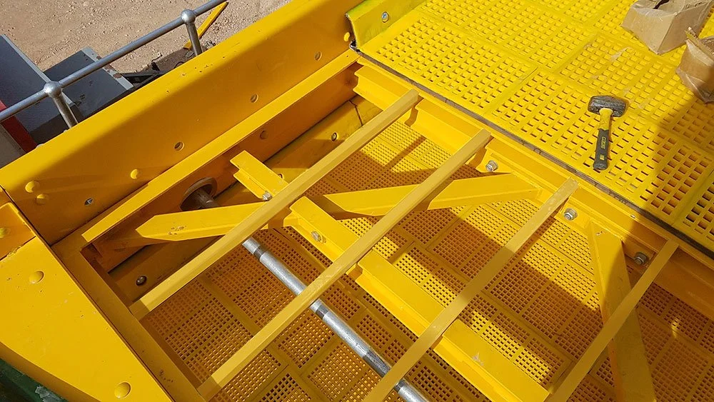 Close-up of a yellow metal machinery part with a grid-patterned surface and metal rods, with a screw and hammer placed on the surface.