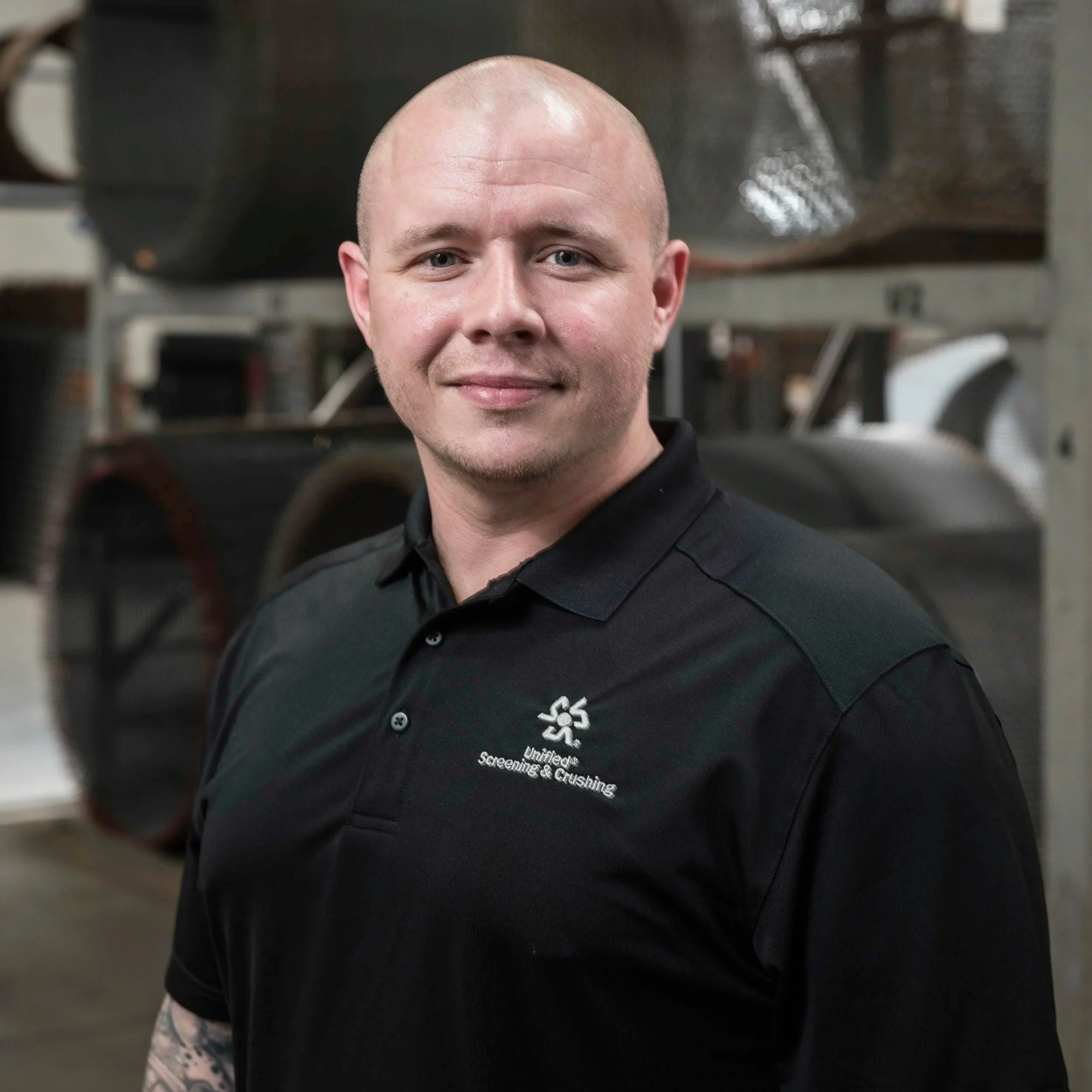 A young man with a shaved head, wearing a black shirt with white text on it. The background shows gym equipment, indicating he is in a gym or fitness center.