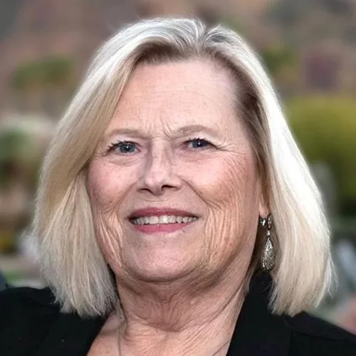 Close-up of an older woman with shoulder-length blonde hair, smiling, wearing earrings and a black top, outdoors with blurred greenery in the background.
