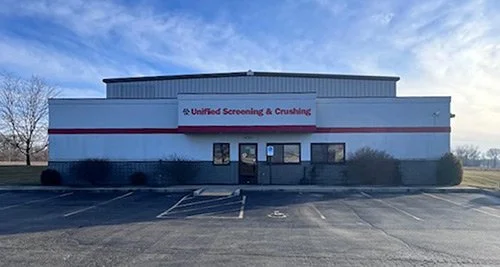 Empty parking lot in front of a commercial building with a sign that reads 'Unified Screening & Crushing' under a partly cloudy sky.